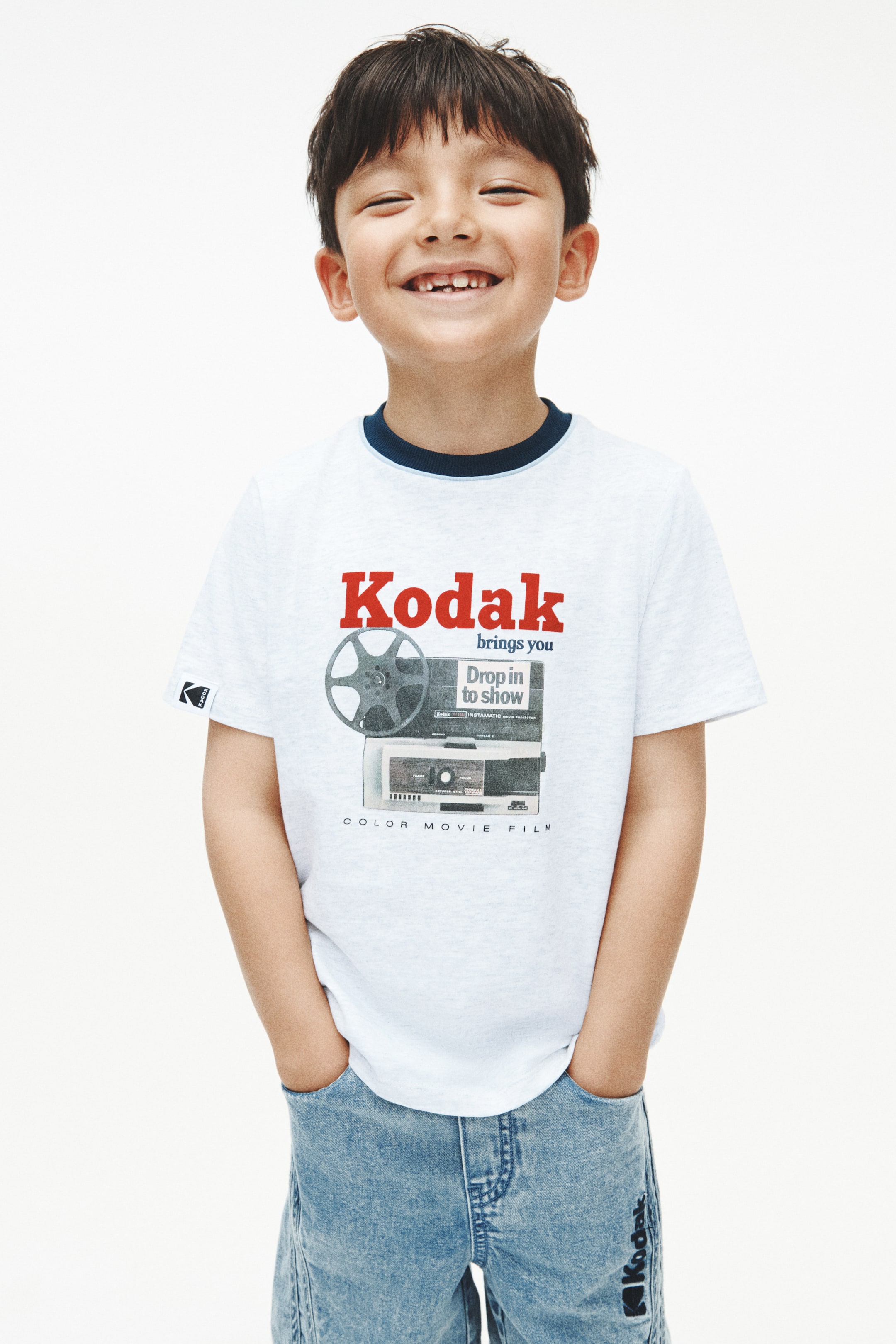 이미지 확대 보기: 연한 회색 반팔 티셔츠는 필름 릴과 프로젝터의 일러스트 위에 "Kodak brings you"라는 빨간 로고가 특징이며, 네이비 블루 링거 넥라인과 왼쪽 소매에 작은 Kodak 로고가 있습니다. 연한 파란색 데님 반바지도 착용하고 있으며, 오른쪽 하단 다리에 세로로 "Kodak" 로고가 표시되어 있습니다.