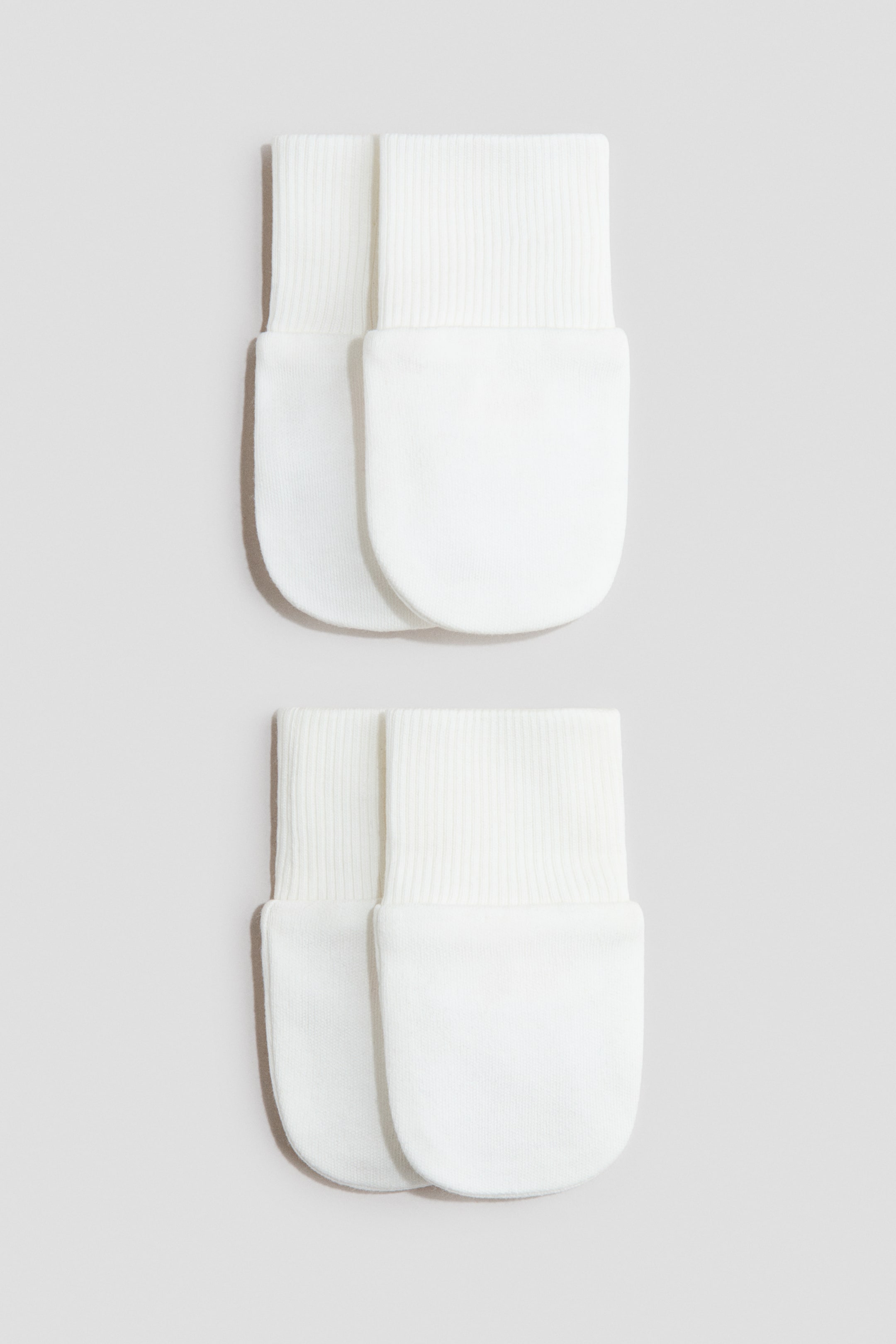 View larger image: Two pairs of white thumbless scratch mittens are laid out flat, displaying their front. Each mitten showcases a smooth jersey body with ribbed cuffs.