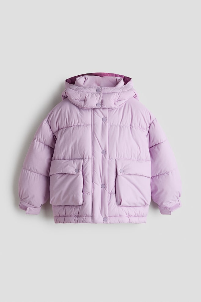 This front-facing light purple puffer jacket is displaying horizontal quilting, a high-neck hooded collar with a contrasting dark purple lining, and a central snap-button closure. It is featuring two large front utility pockets with snap-flap closures and adjustable cuffs on the sleeves. - 1