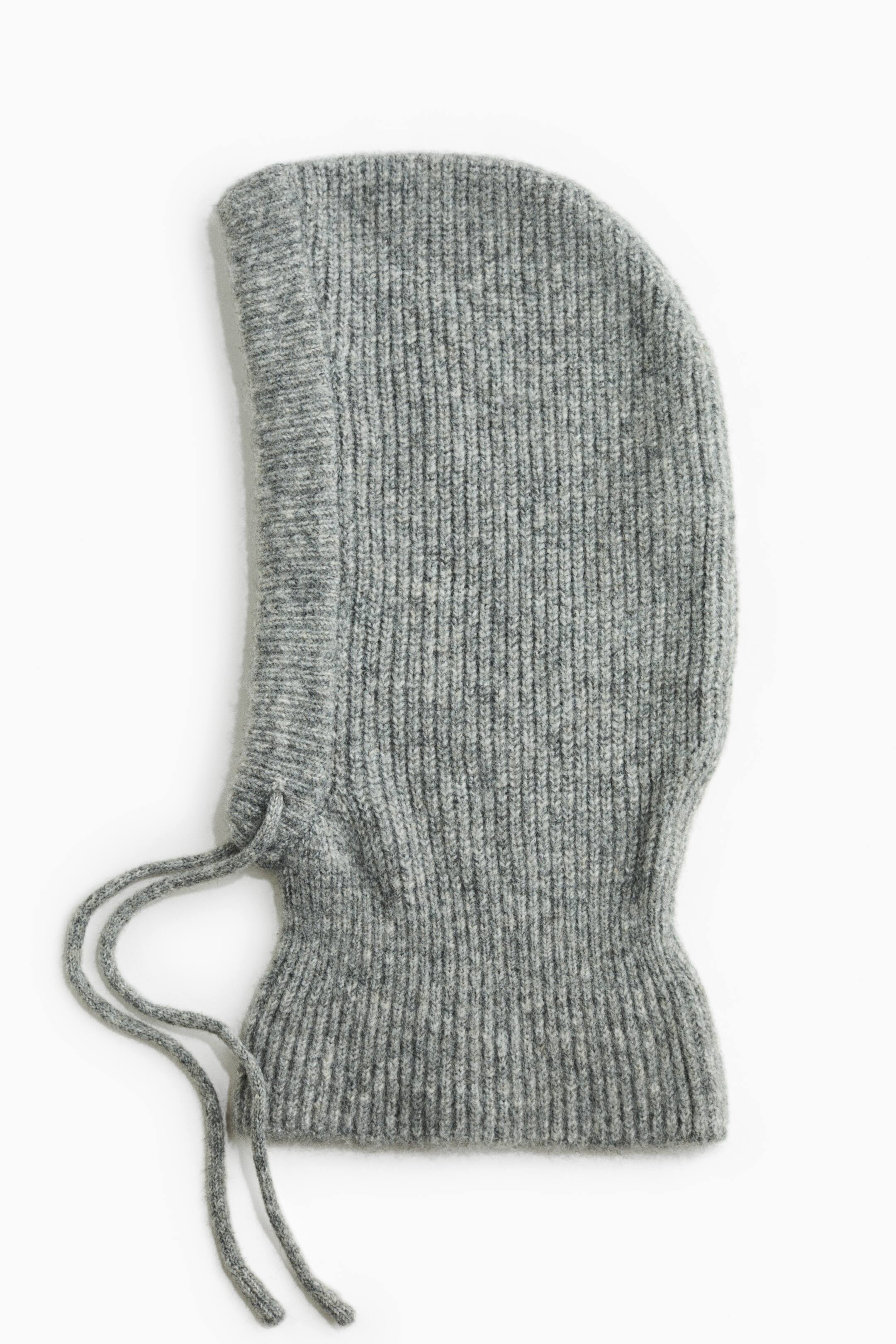 View larger image: A heather grey knitted balaclava is shown in side profile, facing left. The hood features a vertical ribbed-knit construction, a snug ribbed neckband, and two matching knit drawstrings extending from the lower front opening.