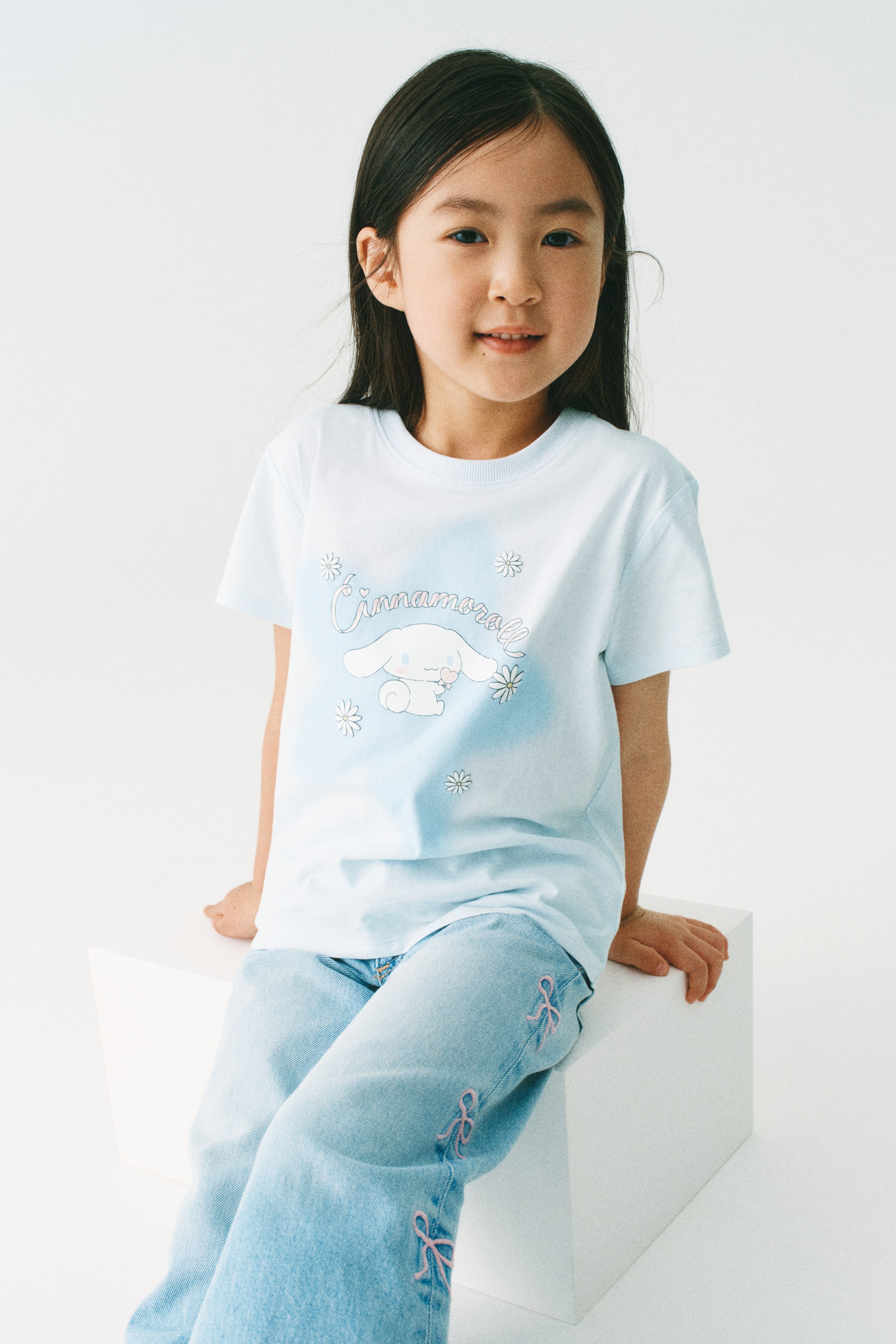 View larger image: A light blue short-sleeved t-shirt is showcased from the front, featuring a round, ribbed neckline and a subtle light blue gradient design. The front displays a white Cinnamoroll character graphic, along with "Cinnamoroll" text and small flower designs.