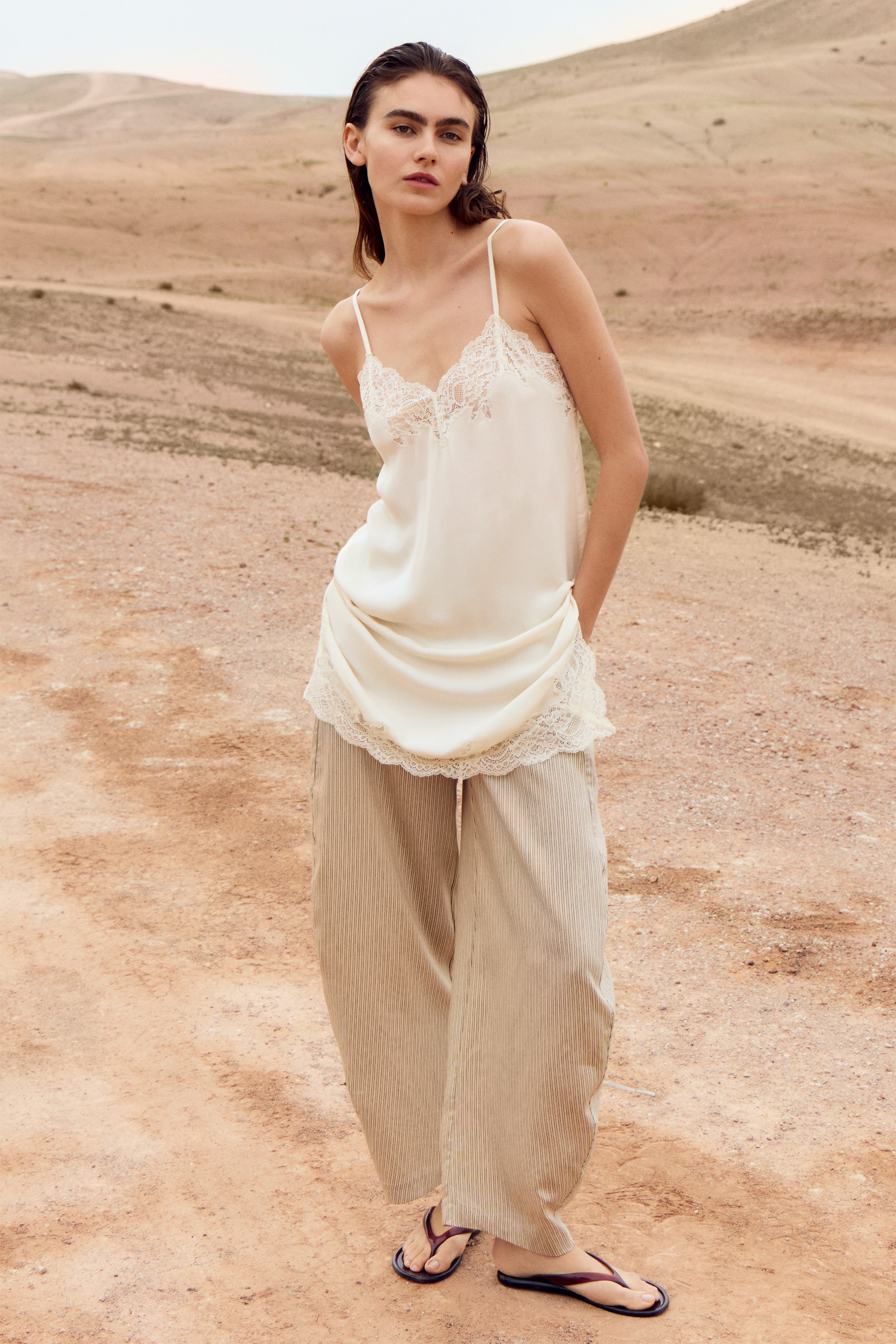 View larger image: An ivory satin camisole with thin straps and delicate lace trim along the V-neckline and hem is worn, draping loosely over full-length, wide-leg beige pants with fine vertical pleats. Dark thong-style flip-flops complete the look.