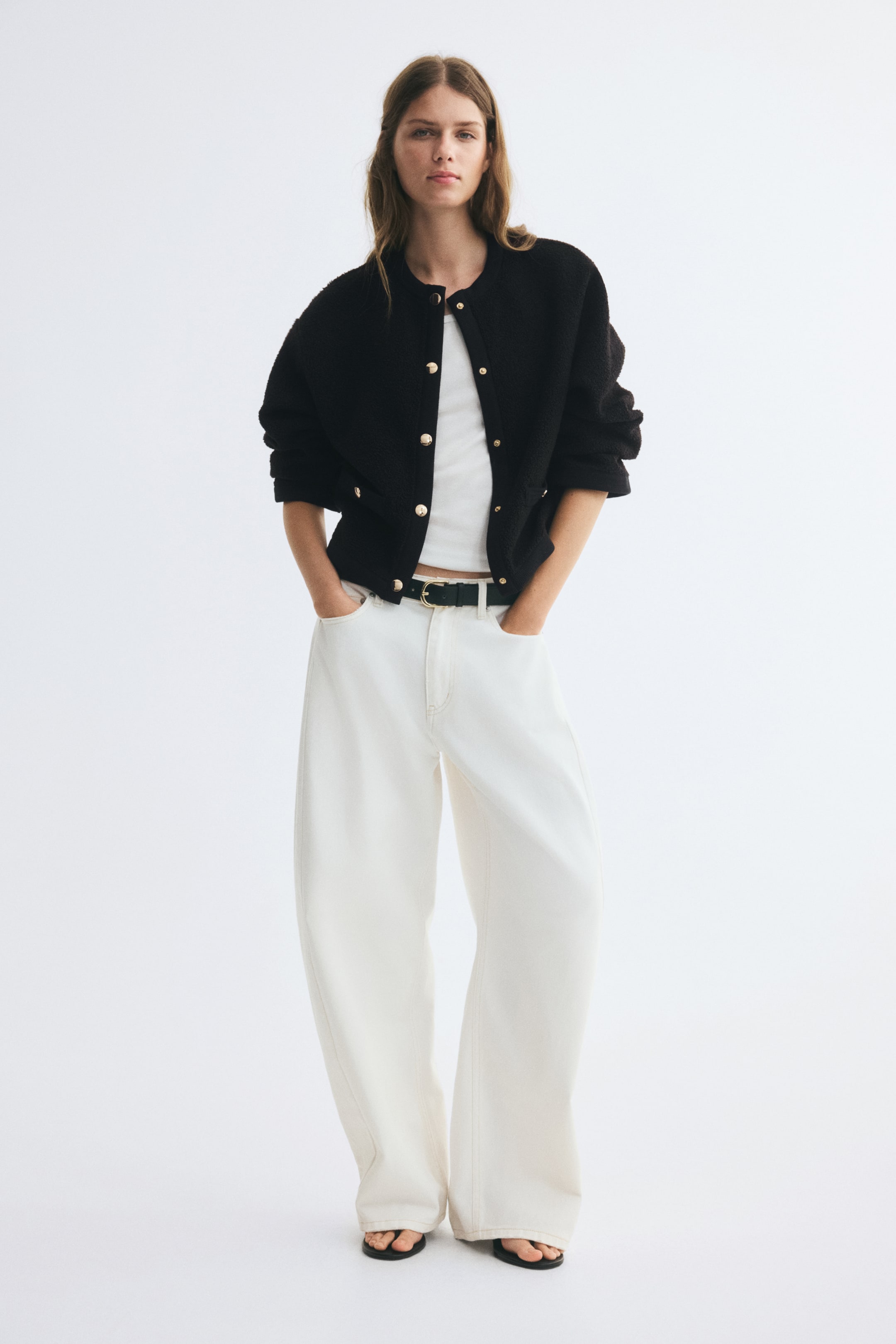 View larger image: A black relaxed-fit teddy jacket with a round neckline, gold-tone metal snap buttons, and mock welt pockets with decorative gold buttons is worn open by the model. The jacket features dropped shoulders and ribbed trims and is layered over a white top and paired with wide-leg white trousers.