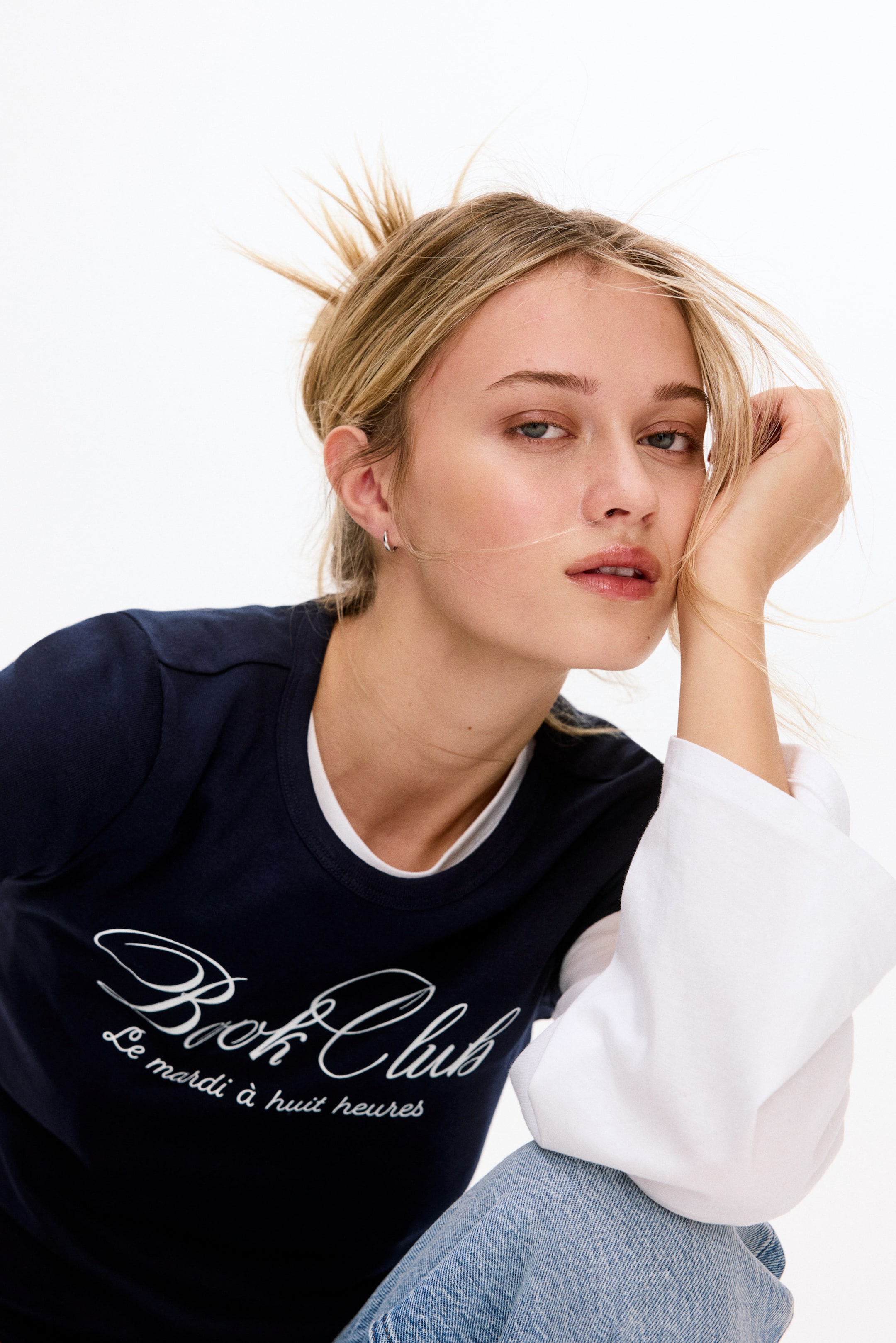 View larger image: A navy blue t-shirt featuring a crew neck and a white "Book Club" graphic, with "Le mardi à huit heures" written below it. It is worn layered over a white long-sleeved top, with the white fabric visibly extending from the neckline and sleeves.