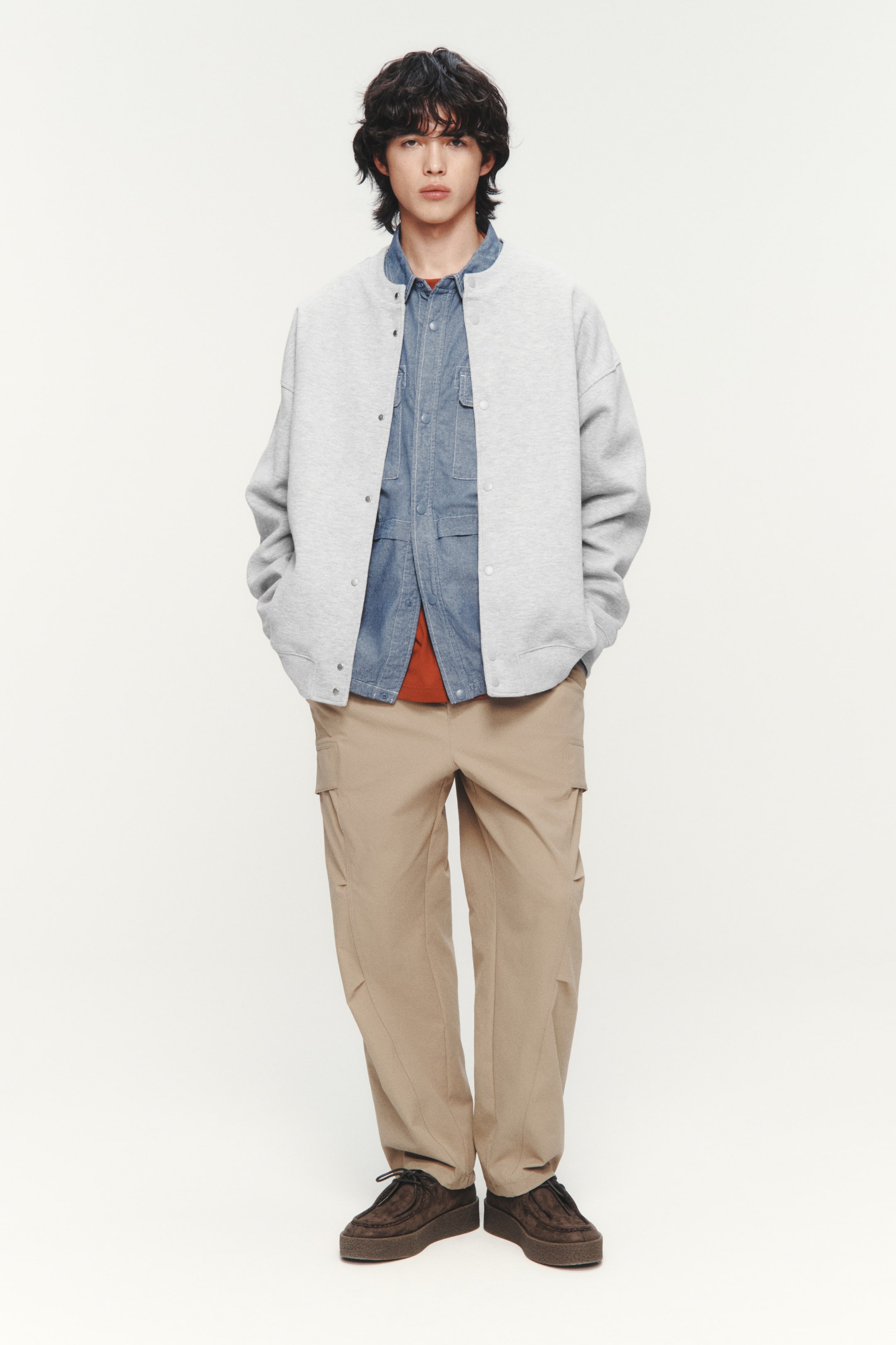 拡大画像を表示: モデルはライトグレーのジャージーカーディガンを着用しています。ラウンドネックラインにはリブトリムが施され、前面にはプレススタッドが並び、ドロップショルダー、長袖、ウェルトフロントポケット、袖口と裾には幅広のリブトリムが特徴です。このカーディガンはゆったりとしたフィット感があり、ブルーデニムシャツとオレンジ色のTシャツの上に重ねて着られています。モデルはポケットに手を入れています。