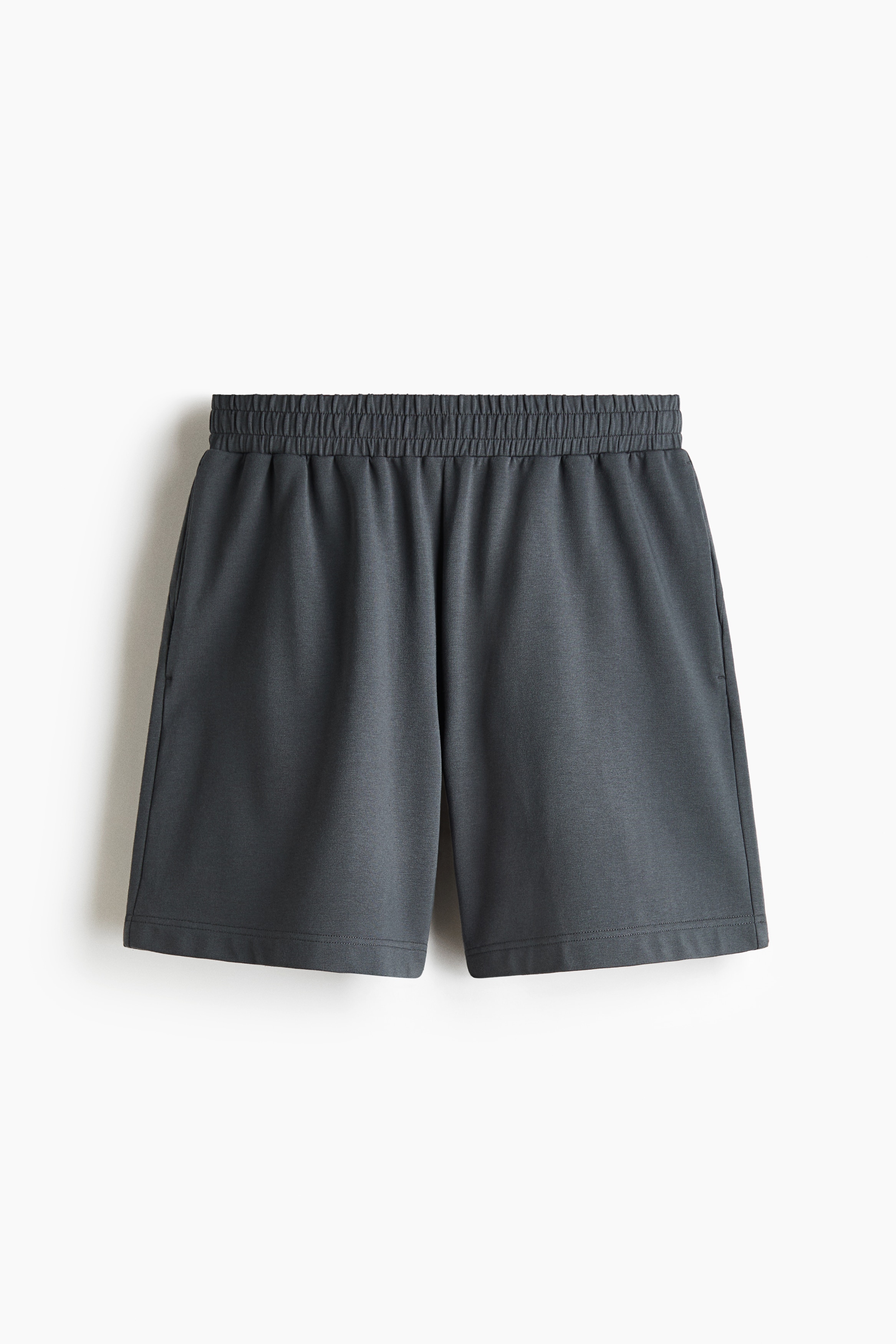 Dark grey sweat shorts featuring an elasticated waistband and a relaxed fit are being worn by the model. The shorts are styled with a matching zip-up jacket and a tucked-in beige top, with the model resting one hand in a side pocket.