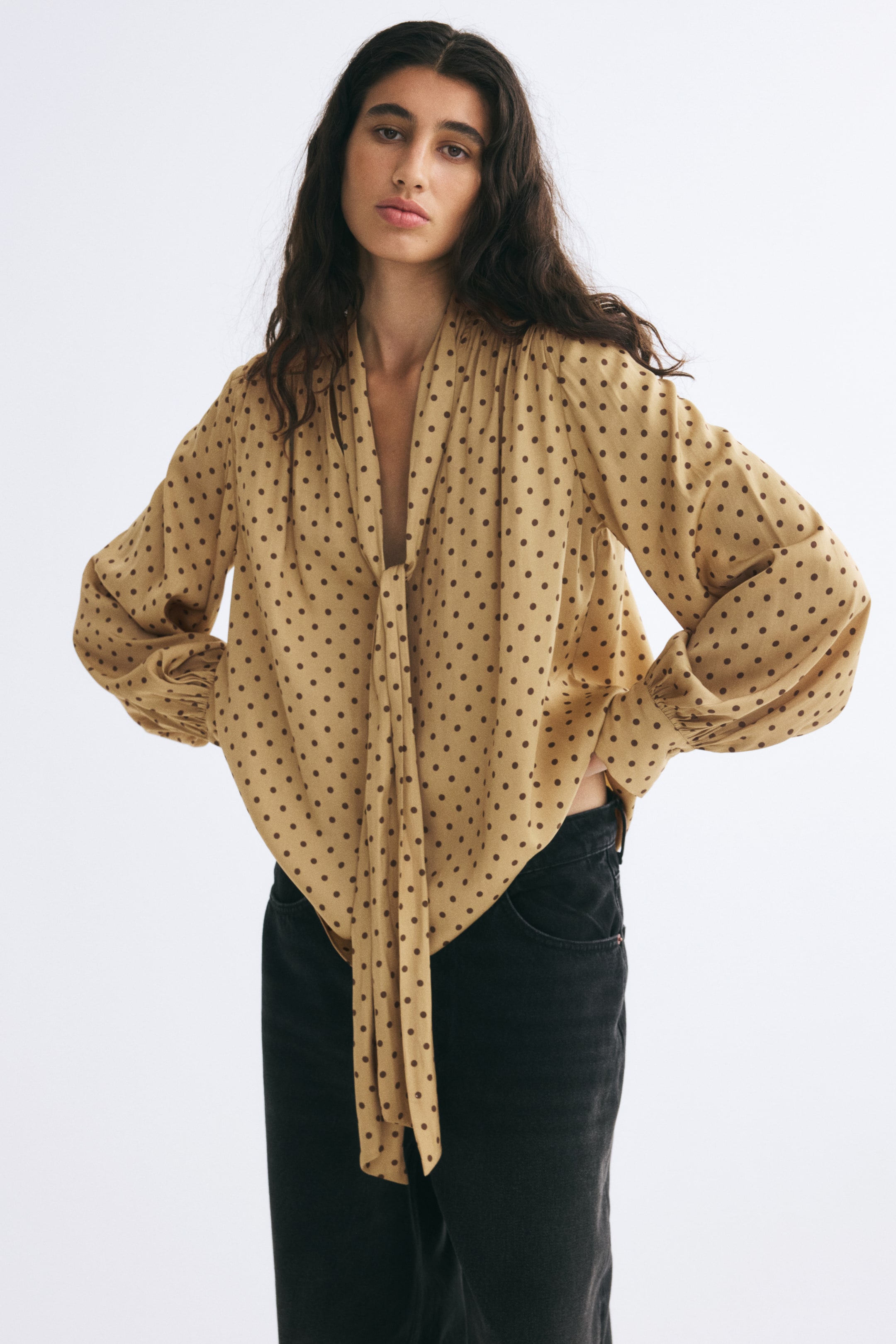 View larger image: The model is wearing an oversized tan polka-dot blouse featuring a V-neck with a long scarf collar tied at the front and gathered detailing for a draped effect. The blouse has voluminous long sleeves with wide button cuffs and is paired with black jeans.