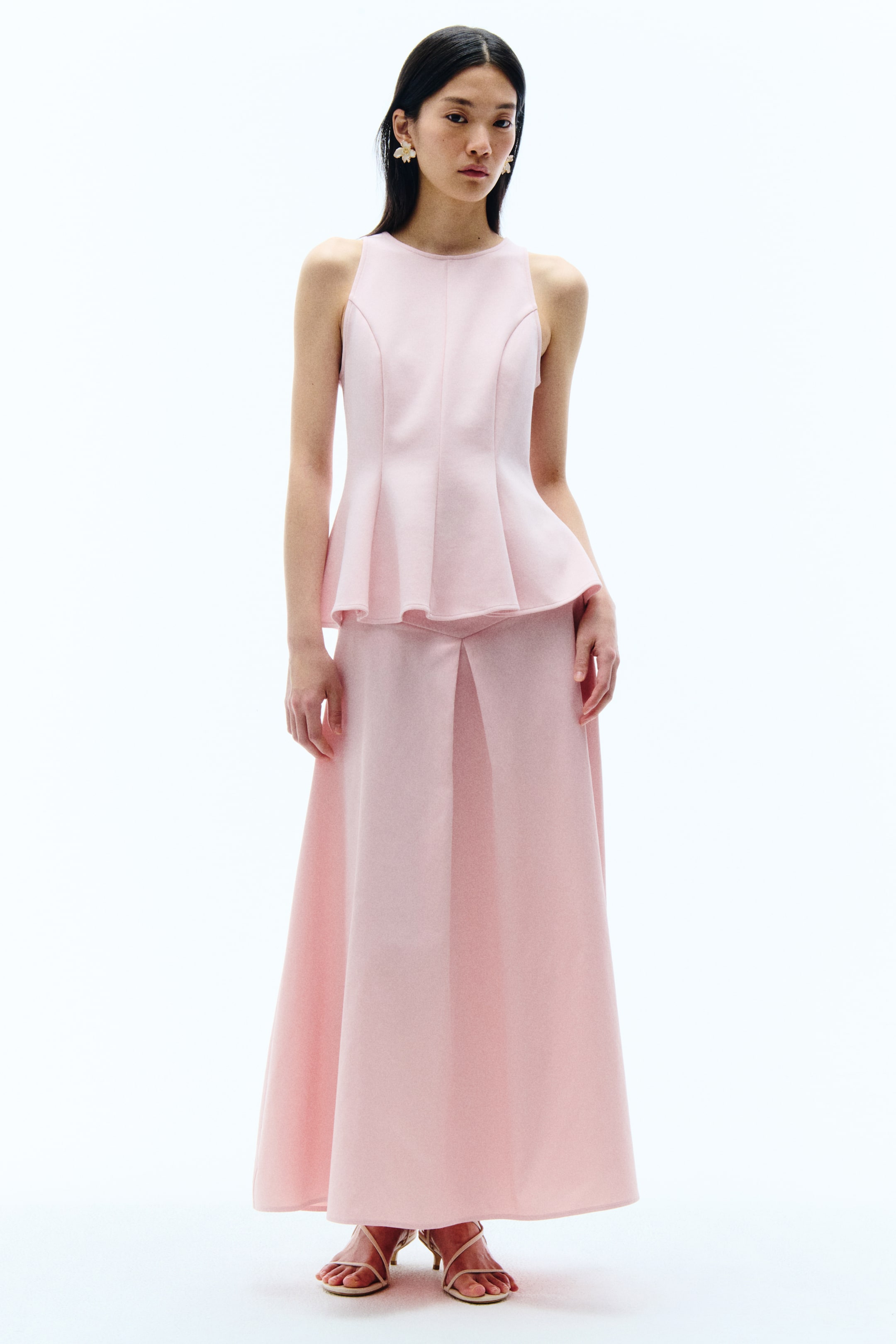 View larger image: A pink two-piece ensemble is shown, featuring a sleeveless peplum top and a matching long skirt. The top has a round neckline and a structured peplum hem, while the skirt comes with a central front pleat.