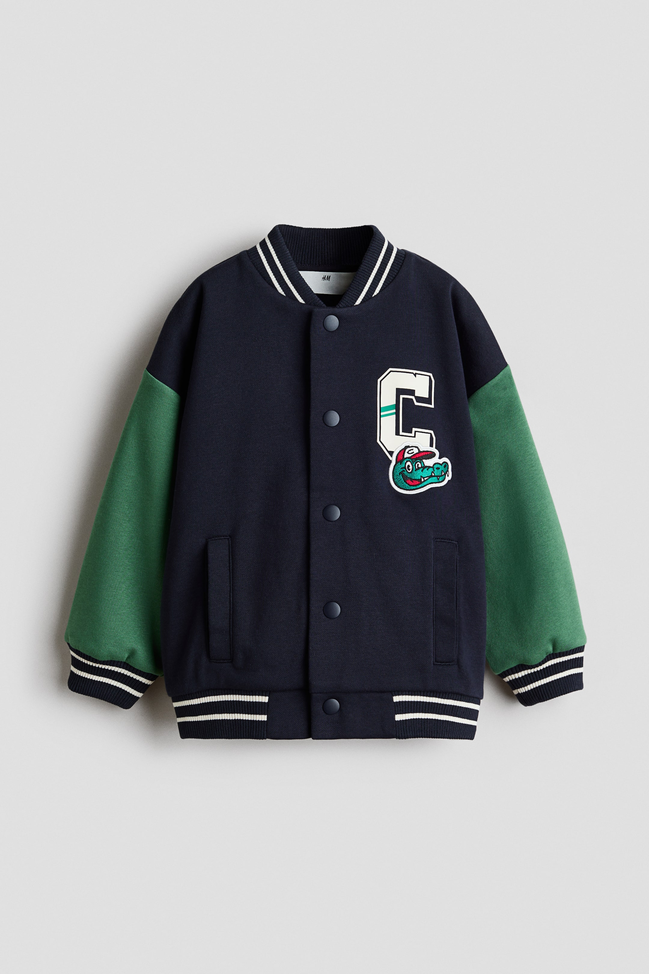 View larger image: A navy blue varsity jacket with green sleeves is displayed from the front, highlighting a ribbed stand-up collar, ribbed cuffs, and a ribbed hem, all accented with white stripes. Snap buttons secure the front, and two welt pockets are visible. A large letter "C" patch featuring a cartoon alligator sporting a red baseball cap embellishes the left chest.