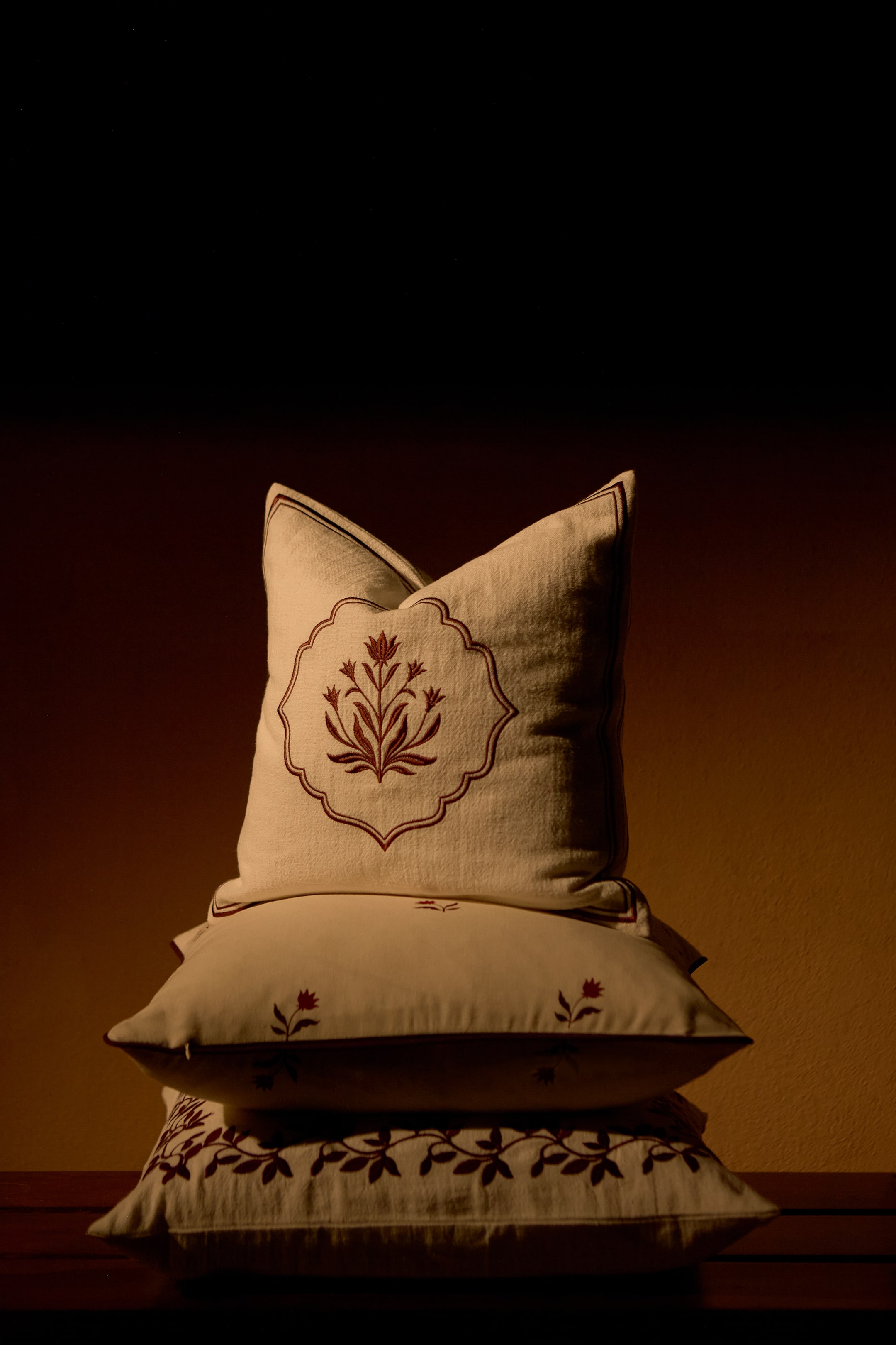 View larger image: A stack of three off-white cotton and linen canvas cushion covers is displaying coordinating dark red embroidered designs from the front. The top cushion is standing upright to showcase a central floral motif enclosed in a decorative frame, while the lower two are resting flat underneath to display their matching botanical patterns.