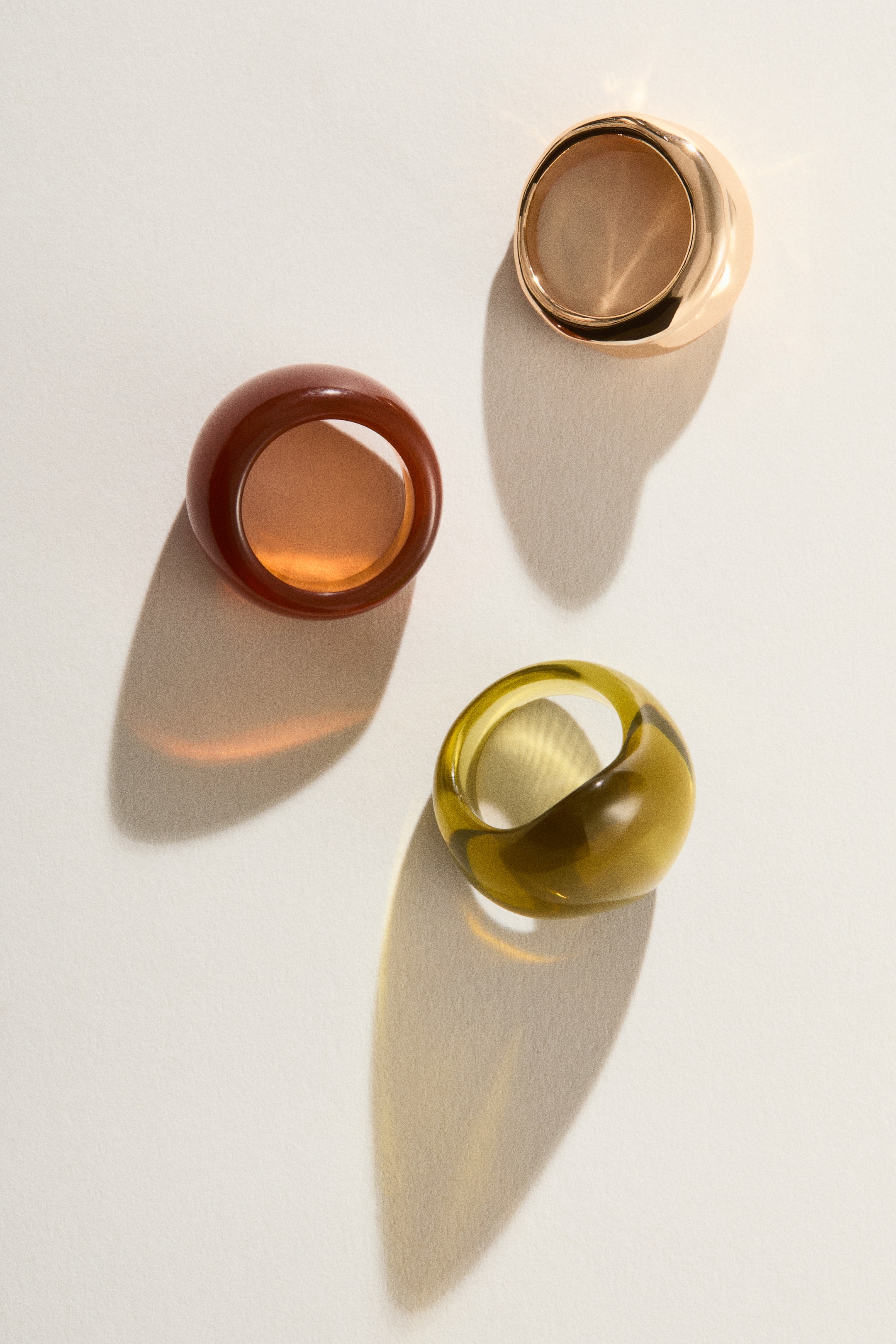 Vis større bilde: Tre kraftige ringer viser ulike design. Den ene ringen er i metall med gulltone og har en blank, ugjennomsiktig finish. De to andre ringene er i gjennomsiktig plast, den ene i en rødbrun farge og den andre i olivengrønn, og alle tre ringene er vinklet for å vise innsiden av ringbåndene.
