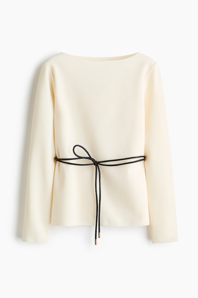 A cream-coloured, loose-fit top is shown from the front, featuring a delicate boat neckline and long sleeves. A thin, black coated tie belt with metallic tips cinches the waist, tied in a bow at the centre. - 1