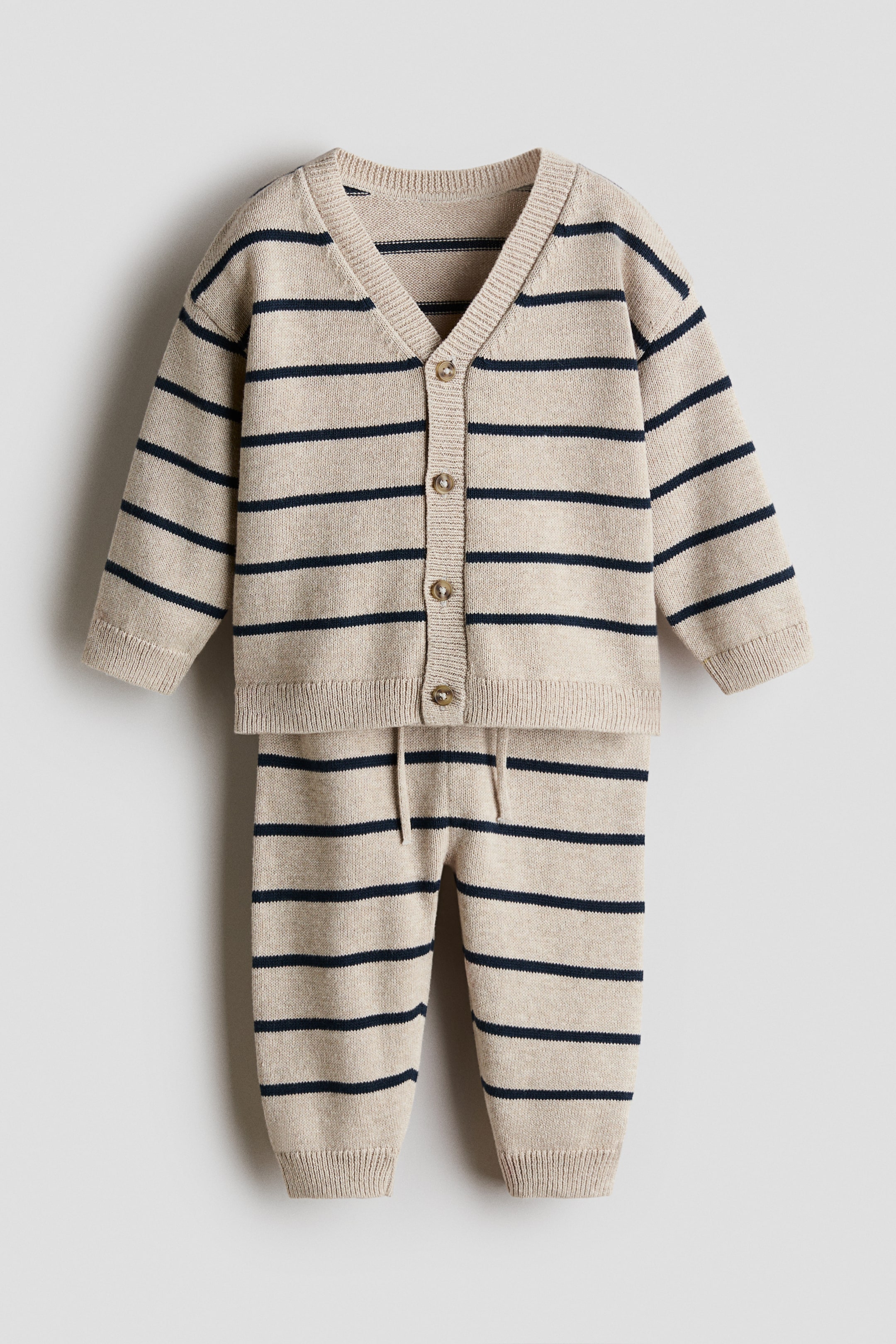 View larger image: A two-piece knitted outfit, consisting of a cardigan and trousers, is lying flat and displaying a beige base with horizontal navy stripes. The cardigan is featuring a V-neck, long sleeves with ribbed cuffs, and a button-front closure with five brown buttons, while the trousers are showing a drawstring waist and ribbed ankle cuffs.