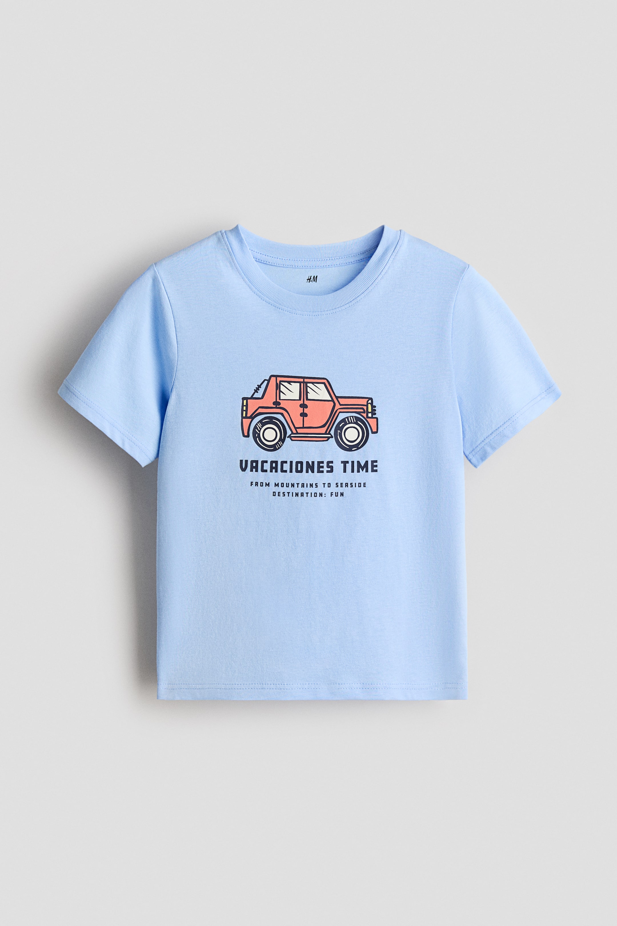 Xem ảnh lớn hơn: Áo thun cotton jersey màu xanh nhạt được trình bày nhìn phía trước, có viền rib quanh cổ, vai rơi và gấu cắt thẳng. Họa tiết in trên ngực mô tả một chiếc xe địa hình màu cam nằm phía trên dòng chữ "VACACIONES TIME" và các chữ nhỏ hơn.