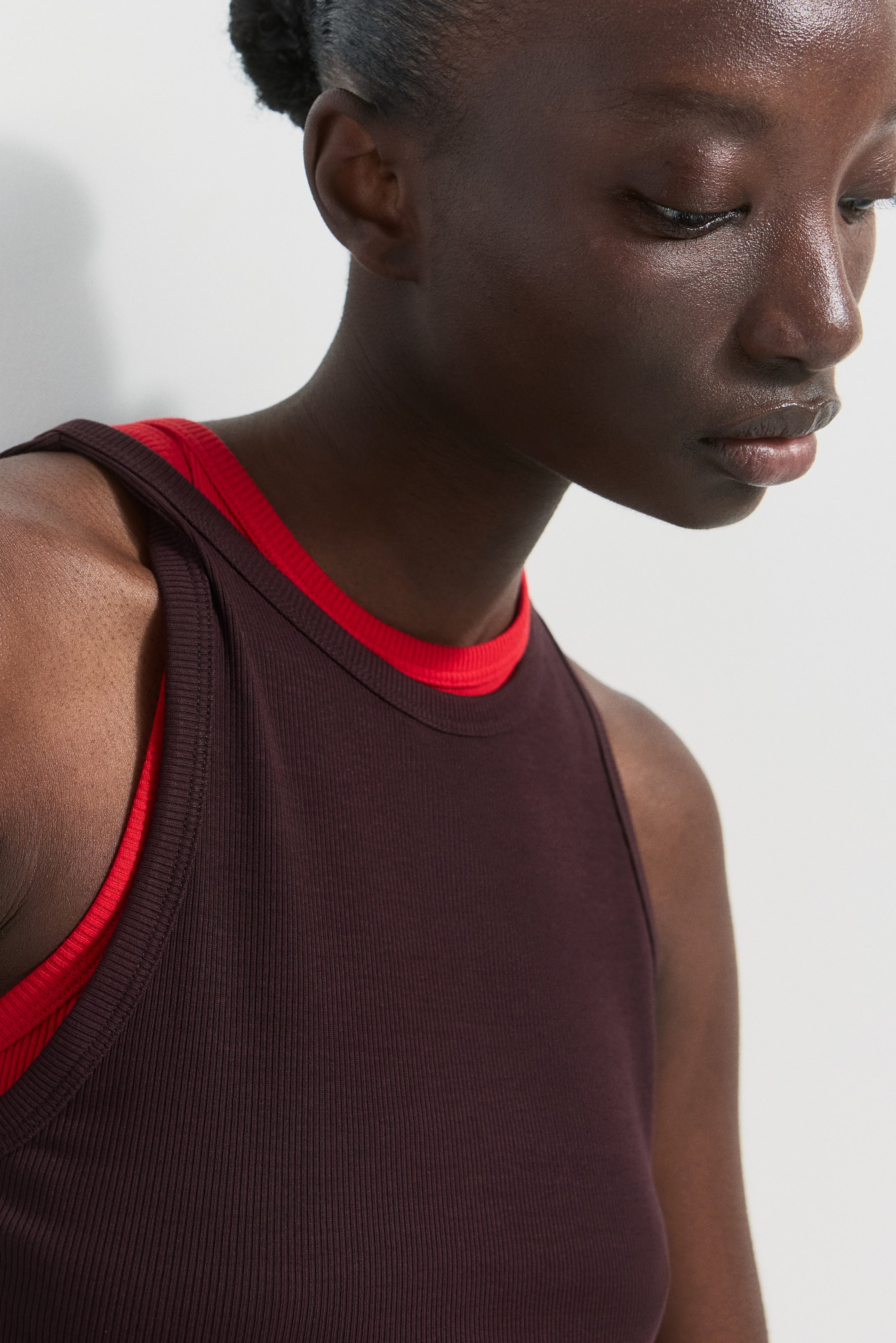 Ver imagen más grande: La modelo lleva puesto un top de tirantes de jersey de algodón acanalado en color marrón oscuro con cuello redondo. Está superpuesto sobre un top rojo, con el cuello rojo visible debajo.