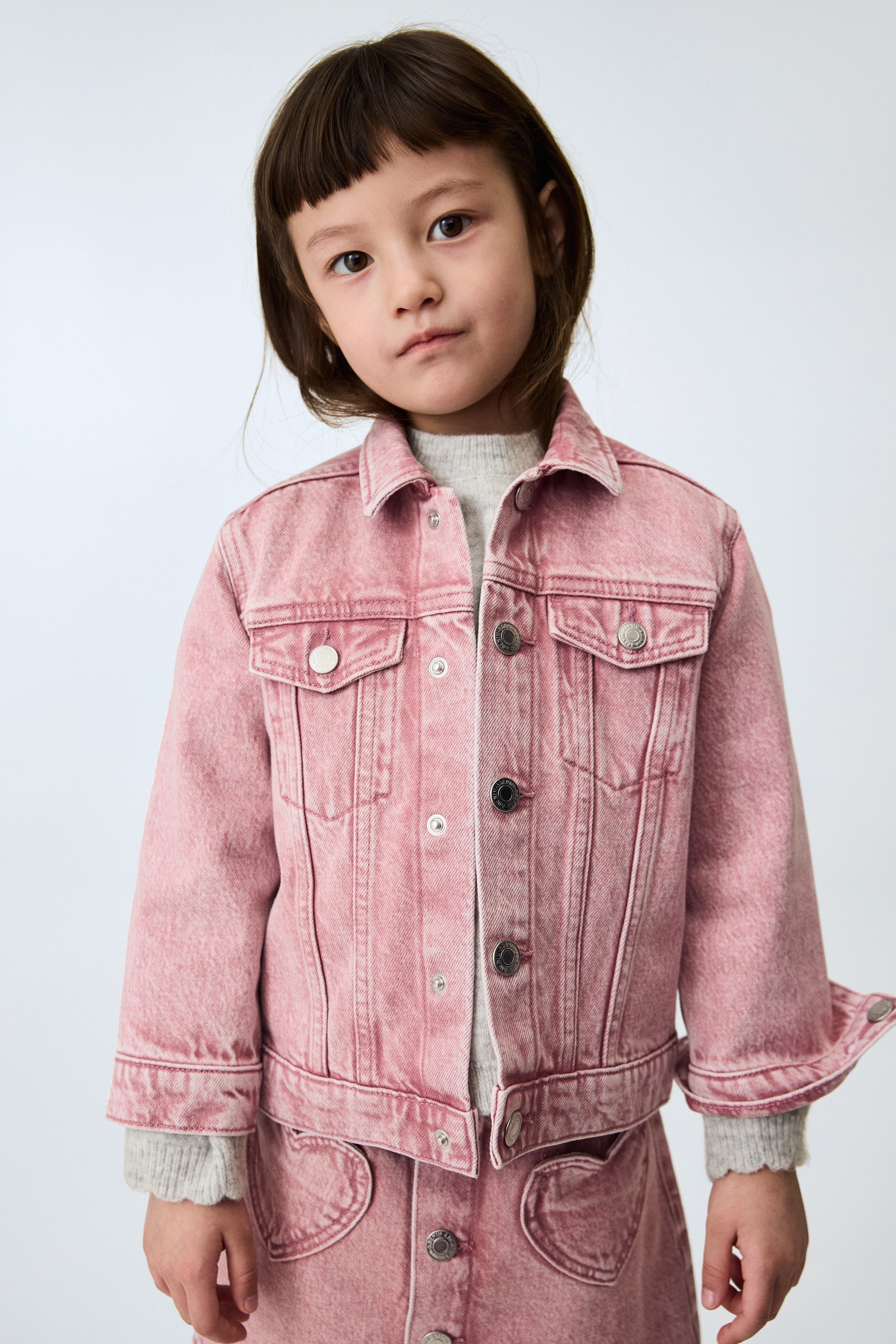 View larger image: A pink washed denim jacket featuring a pointed collar, two flap chest pockets with metal button closures, and a full front button placket. It's layered over a grey ribbed jumper, coordinating with a pink denim skirt showcasing heart-shaped pockets.