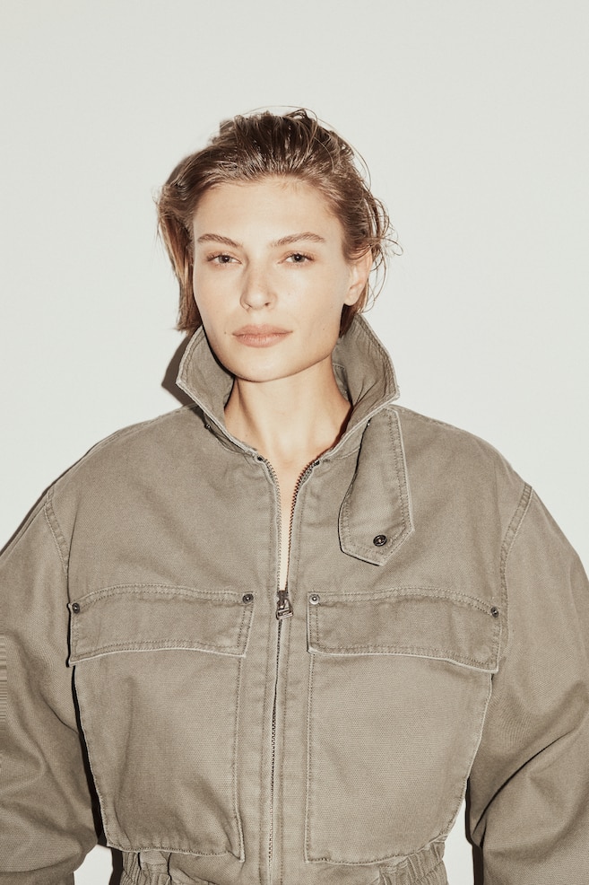 A khaki canvas utility jumpsuit top is shown facing forwards, featuring a standing collar with a snap closure, a full-zip front and two flap chest pockets. An elasticated waistline is visible at the bottom of the garment. - 1