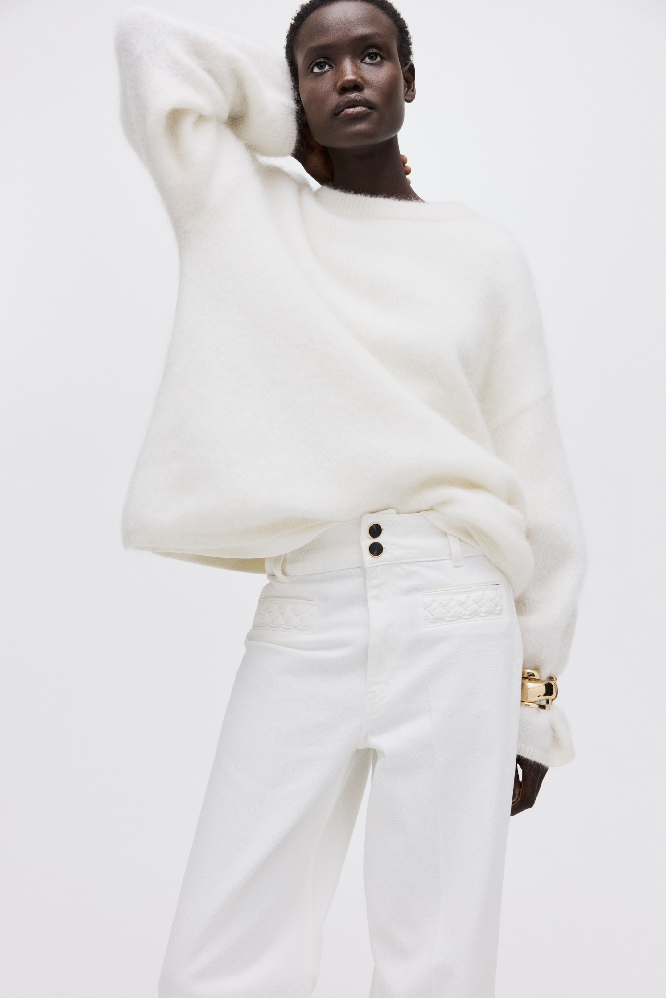 Voir une image plus grande: Un chandail blanc duveteux à col rond est porté, avec des manches longues et volumineuses. Un pantalon blanc à taille haute complète le chandail, légèrement tourné vers l'avant, montrant deux boutons noirs à la ceinture avec des panneaux matelassés décoratifs sur les poches avant de hanches.