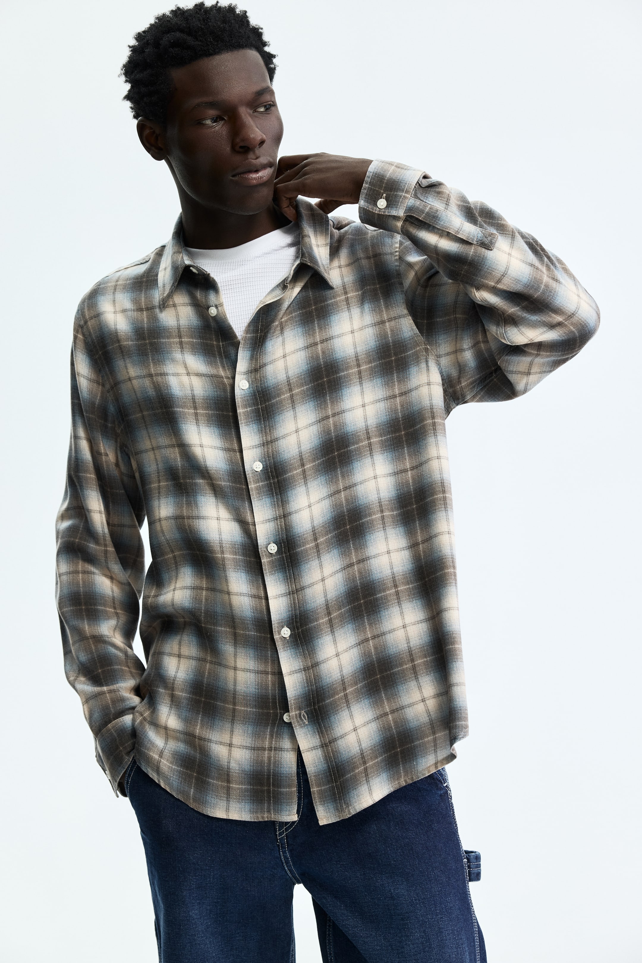 查看大圖: 這款格紋襯衫具有棕色、米色和藍色的漸層色彩，配有長袖、扣袖口和前襟鈕扣閉合設計。從正面角度觀看時，襯衫敞開穿著，搭配白色保暖內衣，突顯出其衣領。