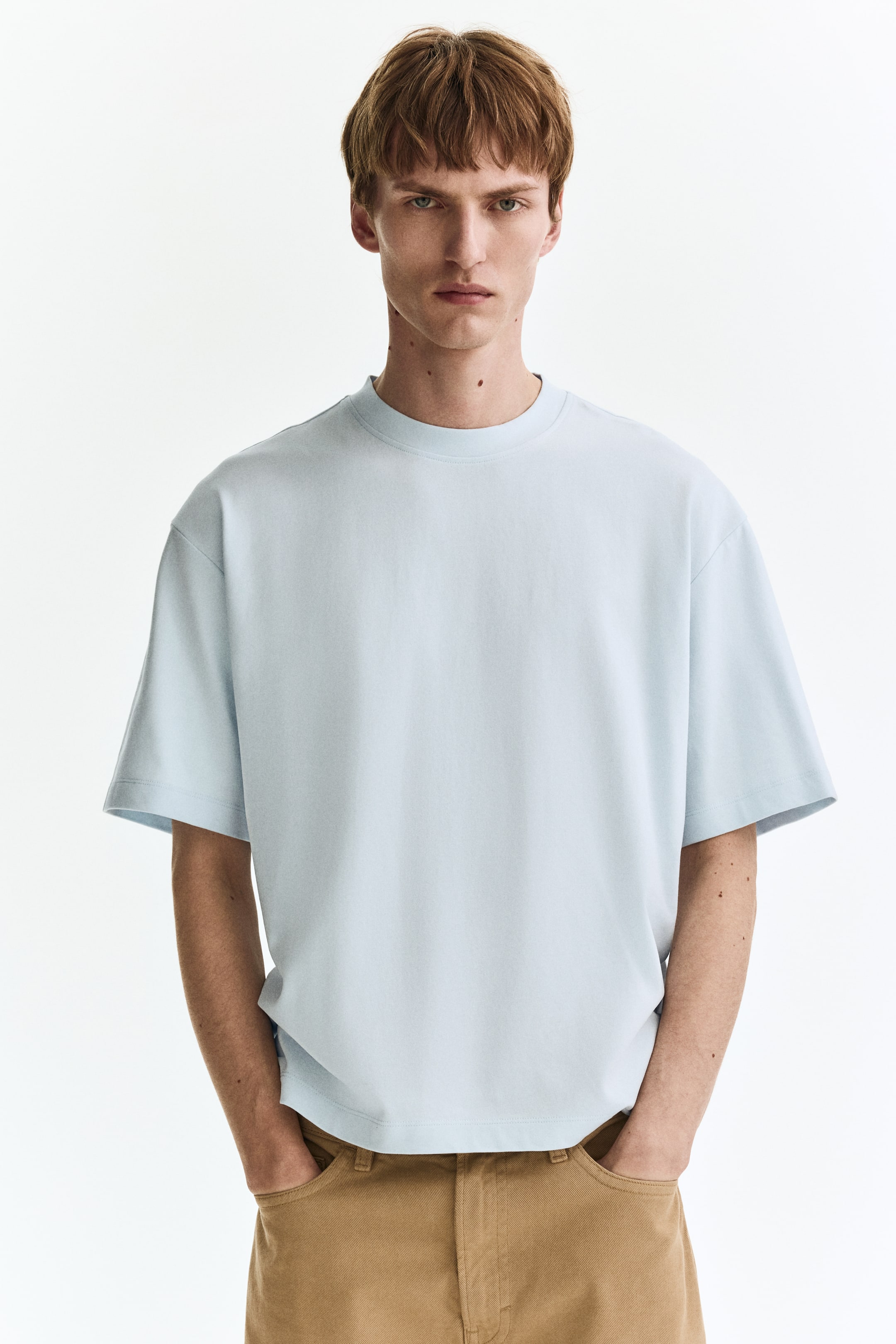Agrandir l'image: Un t-shirt bleu clair est présenté de face, avec un col rond et des manches courtes et larges, et tombe avec une coupe décontractée sur un pantalon marron.