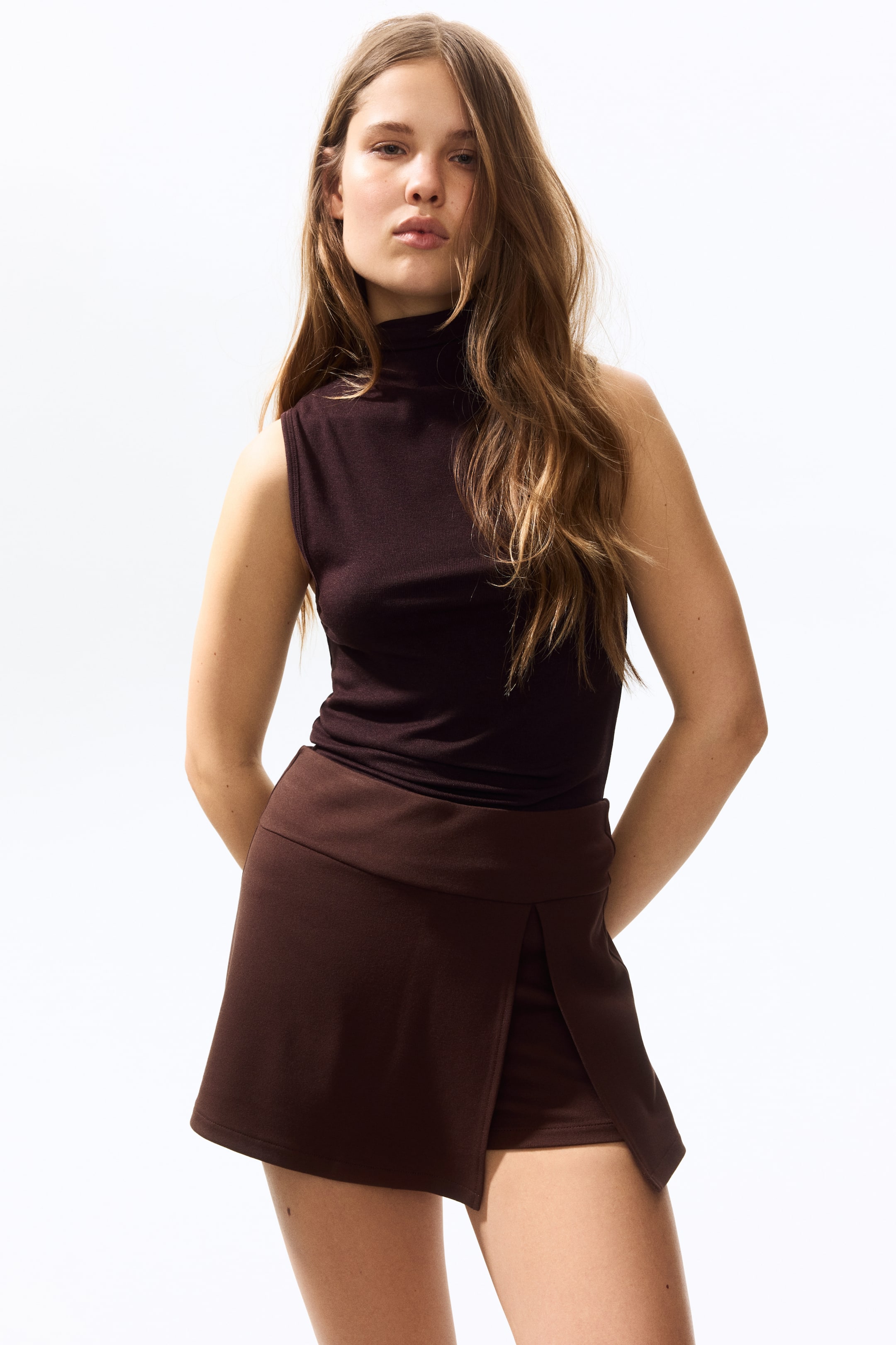 View larger image: A dark brown sleeveless turtleneck top fits snugly and is tucked into a dark brown skort. The skort features a wide waistband and a wrap-front design that creates a side slit.