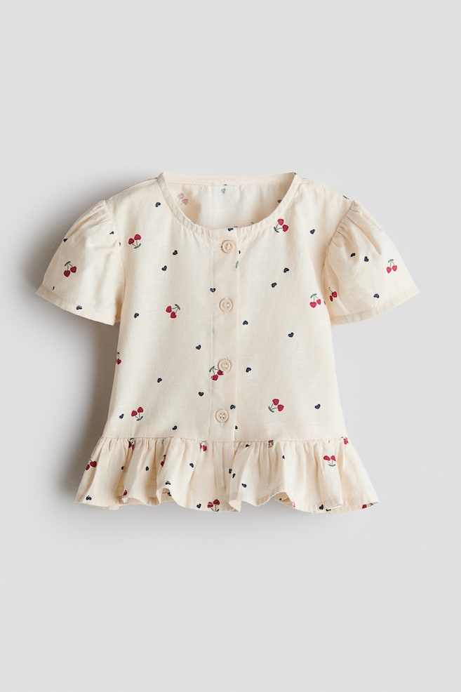 A light-colored blouse in a soft, airy cotton weave is presenting its front, featuring an all-over print of red cherries and small black hearts. It is designed with a round neckline, a button-down front, short puff sleeves, and a ruffled flared hem. - 1