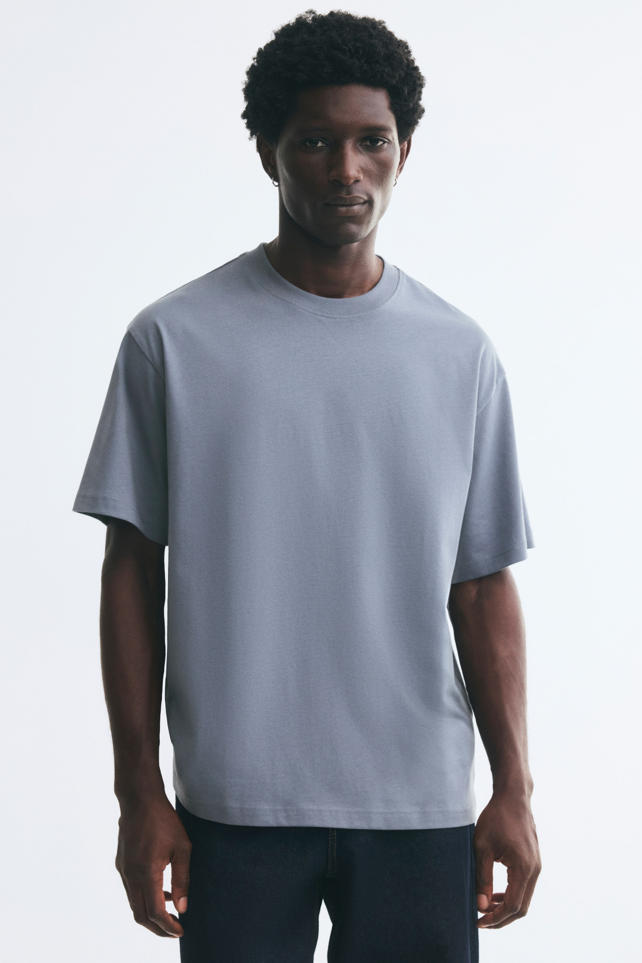 Agrandir l'image: Un t-shirt en jersey de coton bleu-gris clair est porté par le mannequin, avec une coupe décontractée, un col rond bordé de côtes, des épaules tombantes et un ourlet droit. Le t-shirt est assorti à un jean en denim foncé.