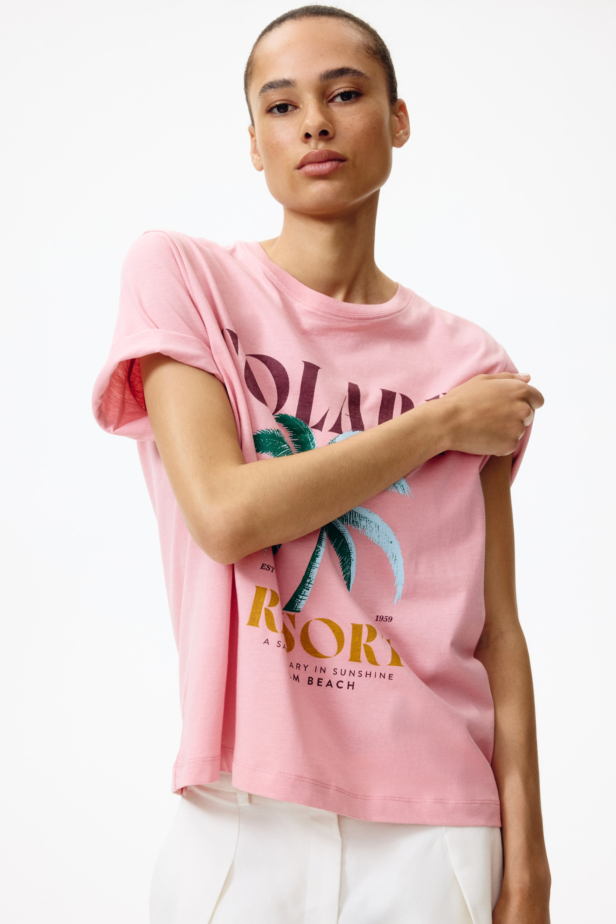 拡大画像を表示: ピンクのクルーネックTシャツは、短いロールアップスリーブと、フロントにヤシの木のグラフィックプリントが施されています。緑とライトブルーの葉が描かれ、部分的に見える「SOLAR」と「RESORT」の文字がダークレッドとマスタードイエローで表現されています。リラックスフィットのTシャツは、白いパンツの上にかかっています。