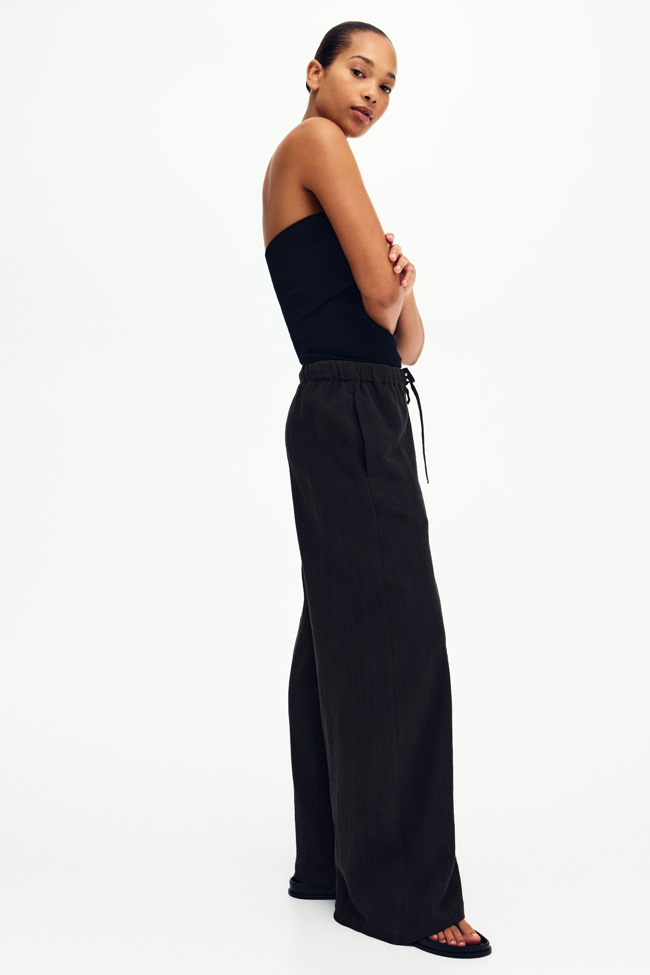 View larger image: Dark, wide-leg trousers featuring an elasticated drawstring waist with a visible tie, side pockets, and a subtly textured fabric, presenting a relaxed fit and wide silhouette from a slight side angle.