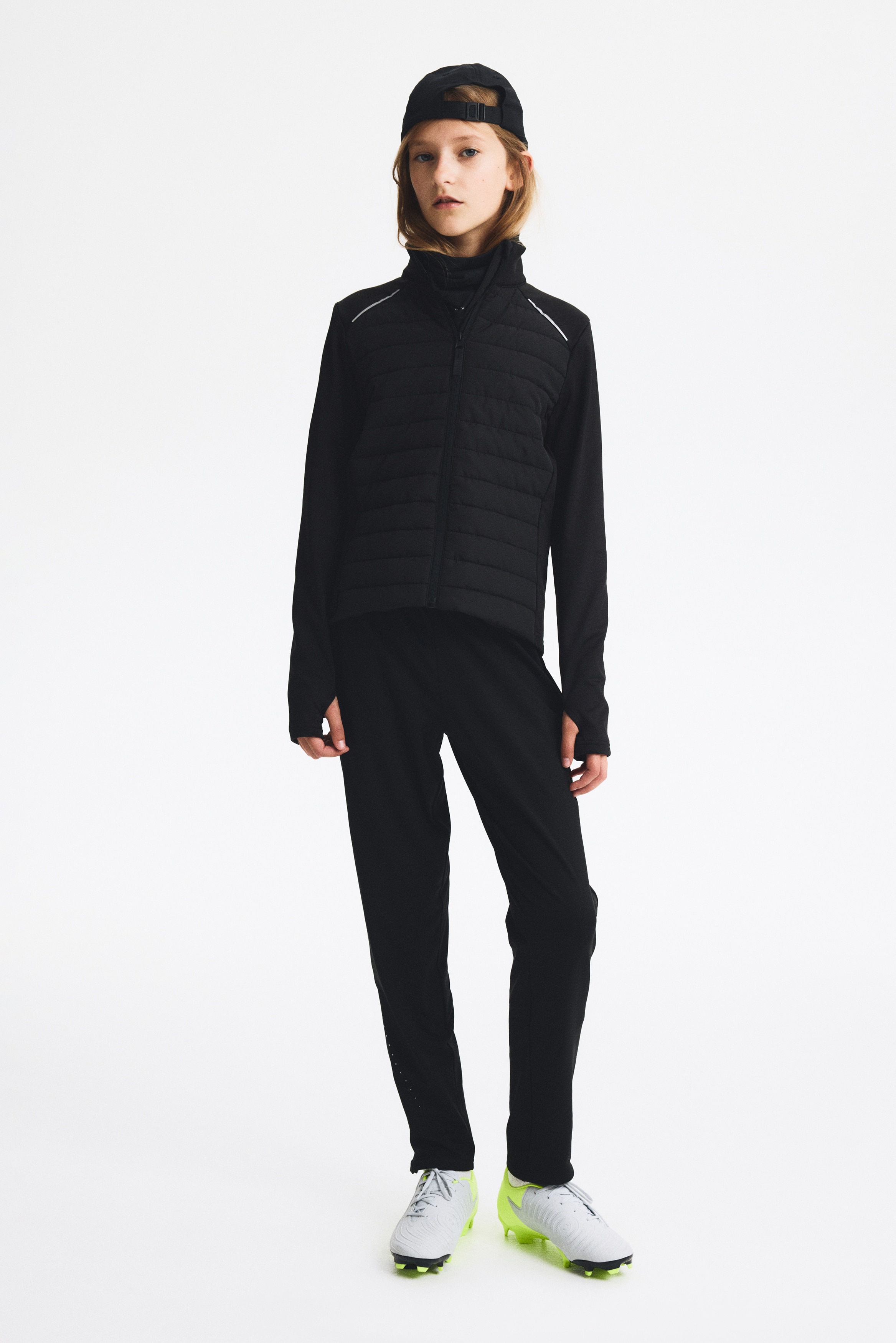 Černá bunda je představována zepředu, má vypolstrovaný a horizontálně prošívaný přední panel, stojatý límec a zapínání na zip po celé délce. Má hladké černé rukávy s otvory pro palce na manžetách a reflexní pruhy jsou viditelné přes horní část hrudníku.