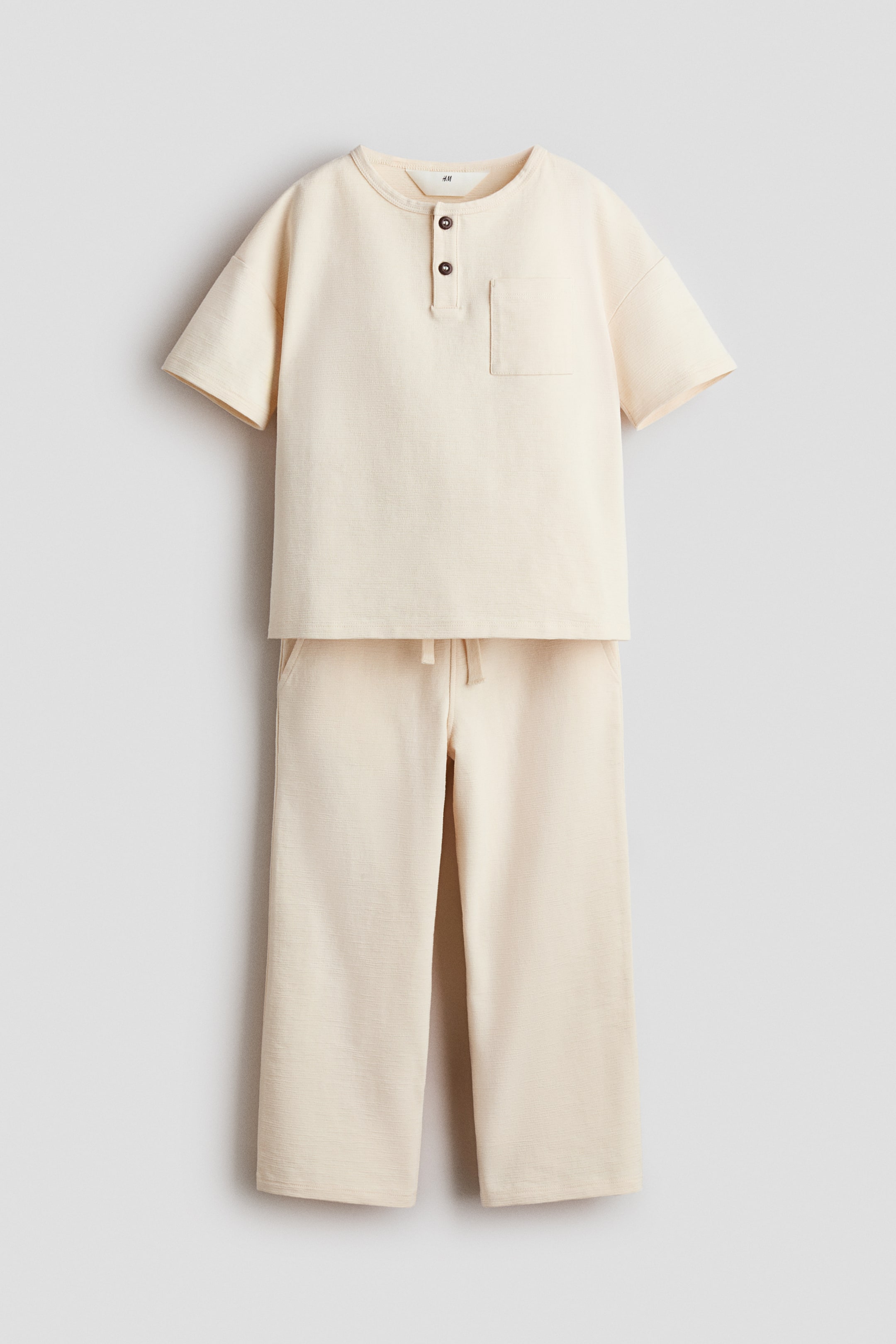Agrandir l'image: Un ensemble deux pièces beige clair se compose d'un haut à manches courtes et d'un pantalon à jambes larges. Le haut présente un col rond, une patte de boutonnage henley à deux boutons et une poche poitrine à gauche, tandis que le pantalon est doté d'une taille à cordon de serrage et de poches latérales.