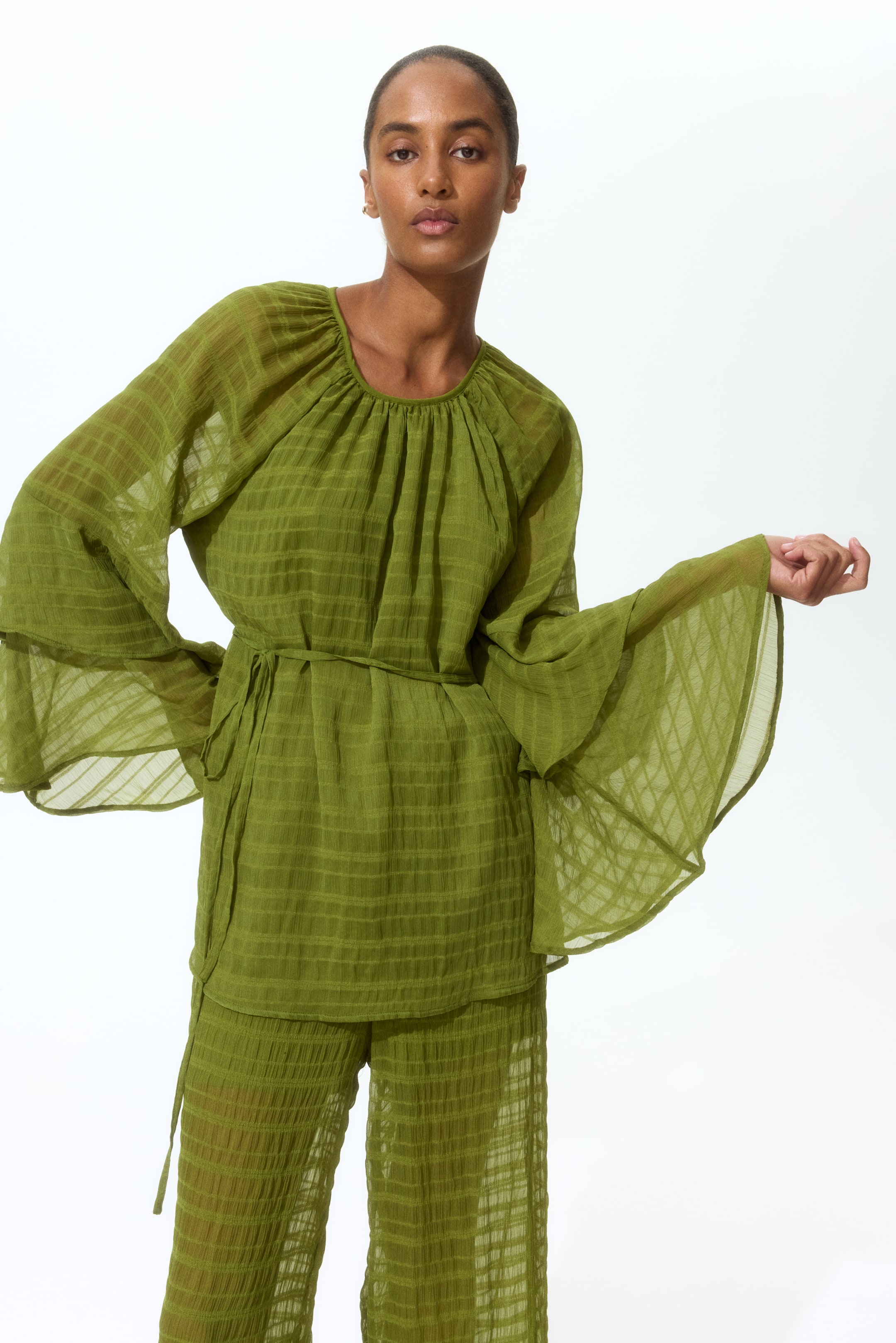 View larger image: A lime green sheer top and matching wide-leg trousers are being showcased from a front-facing angle. The top features a gathered round neckline, tiered bell sleeves, and a self-fabric tie belt that cinches the waist, while the trousers exhibit a loose, flowing fit. Both garments are crafted from a crinkled, grid-patterned fabric.