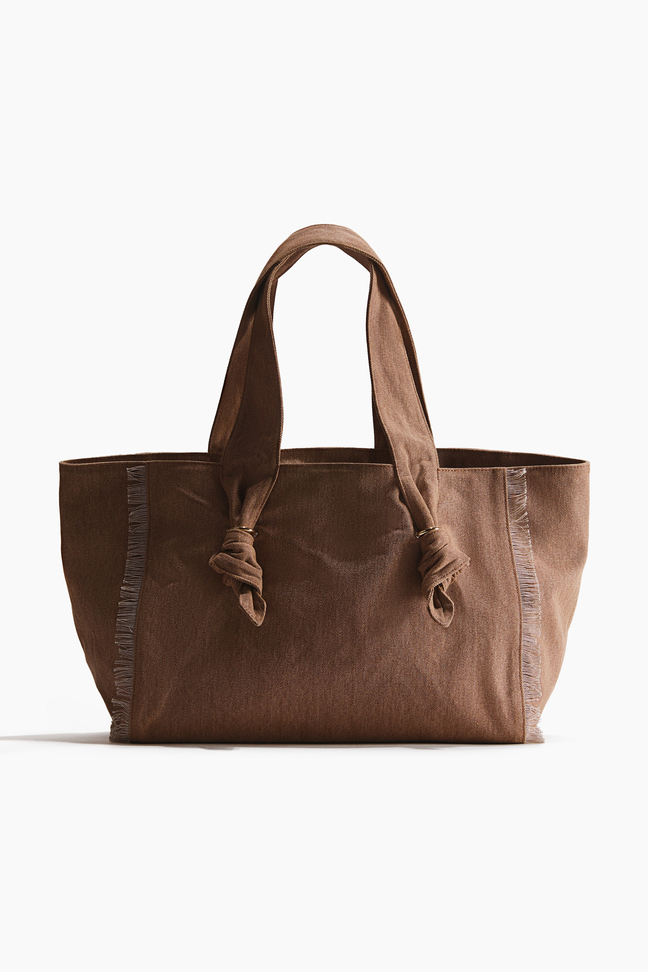 Agrandir l'image: Un sac fourre-tout en tissu marron est orienté vers l'avant, avec deux poignées supérieures nouées avec des anneaux de couleur or, et des détails effilochés verticaux soulignant ses coutures latérales.