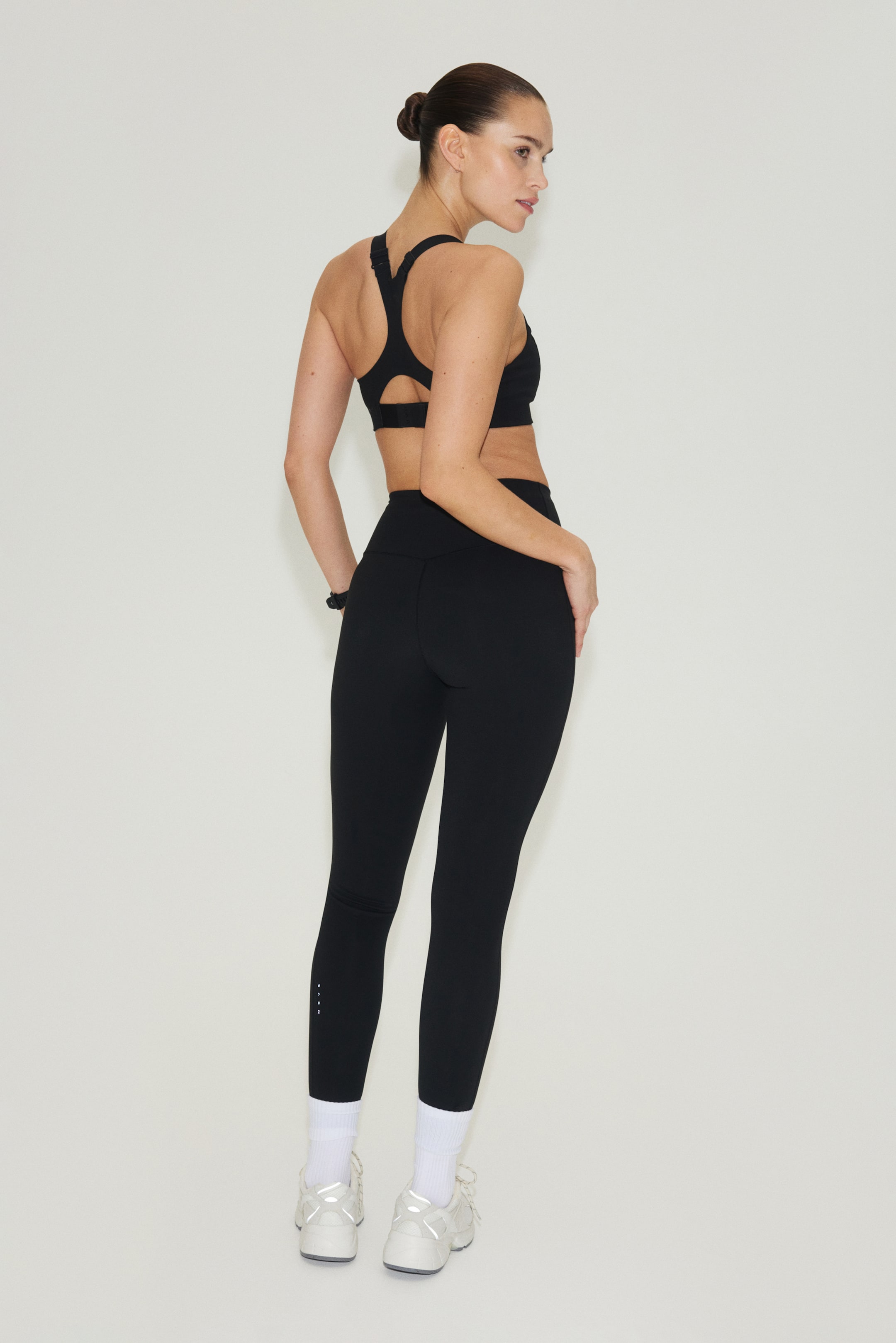 Ingrandisci l'immagine: Un reggiseno sportivo nero con un design a spallina a Y e ritagli viene indossato, visto da un'angolazione posteriore di tre quarti. I leggings neri a vita alta abbinati aderiscono perfettamente, mostrando piccoli punti bianchi vicino alla caviglia sinistra.