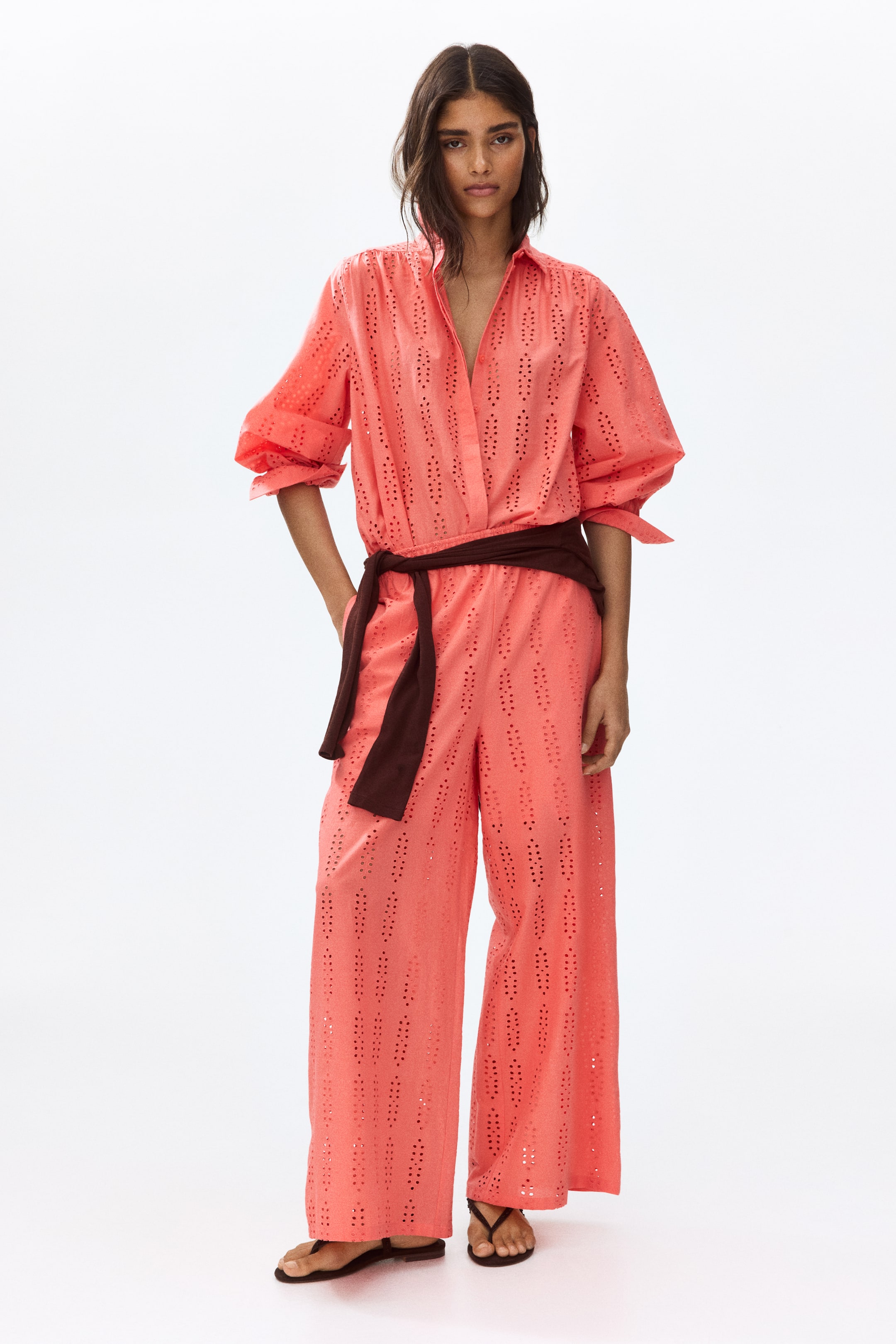 View larger image: The model is donning coral trousers crafted from a light cotton weave with broderie anglaise detailing. These trousers boast wide legs and an elasticated waistband for added comfort. A dark belt cinches the waist, while the model poses elegantly with one hand casually placed in a side pocket.