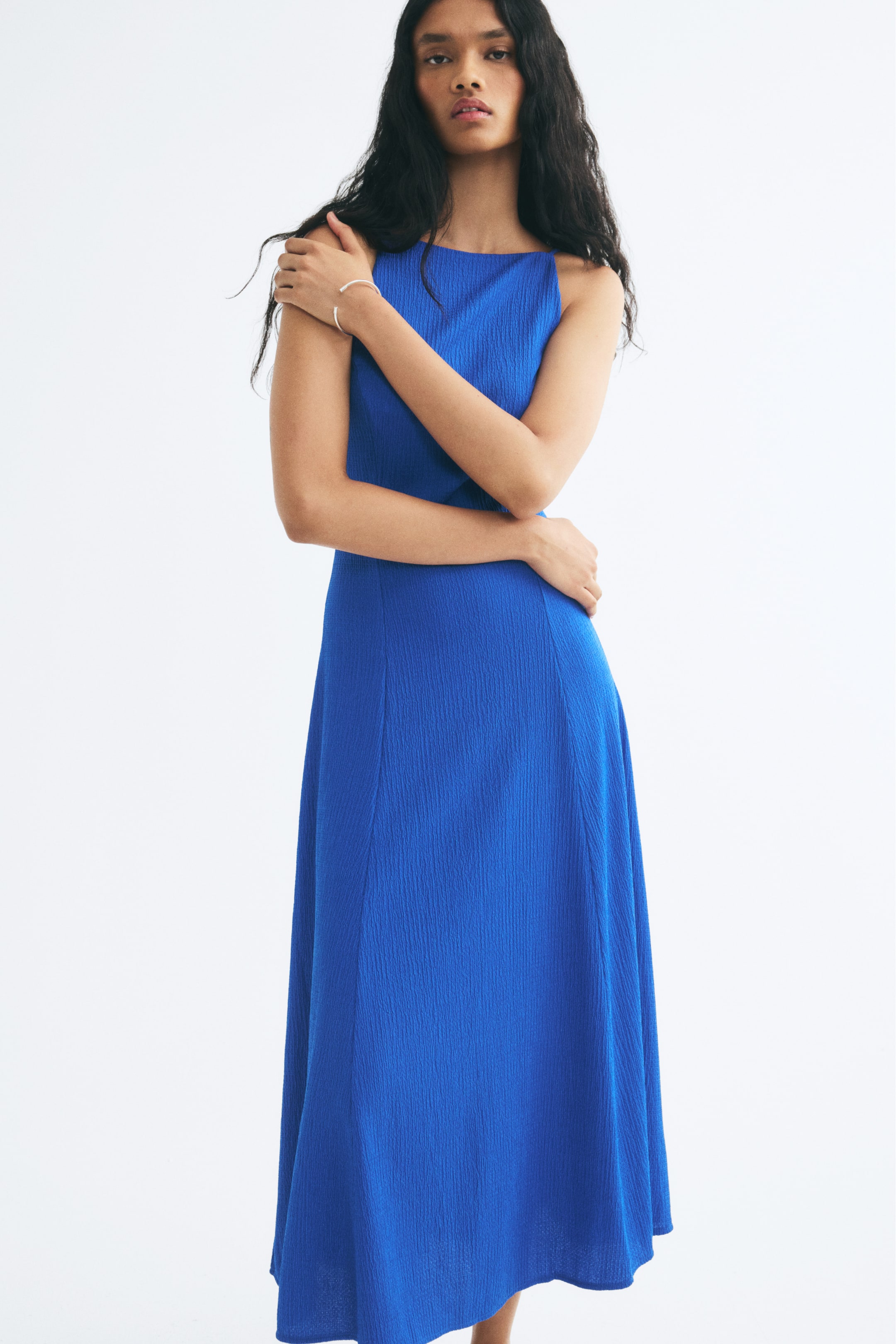 View larger image: A blue crinkled jersey calf-length dress is worn by the model, featuring a square neckline, a fitted bodice with visible vertical seams, and a flared skirt. The model poses with her arms crossed over her chest, highlighting the textured fabric and fitted silhouette.