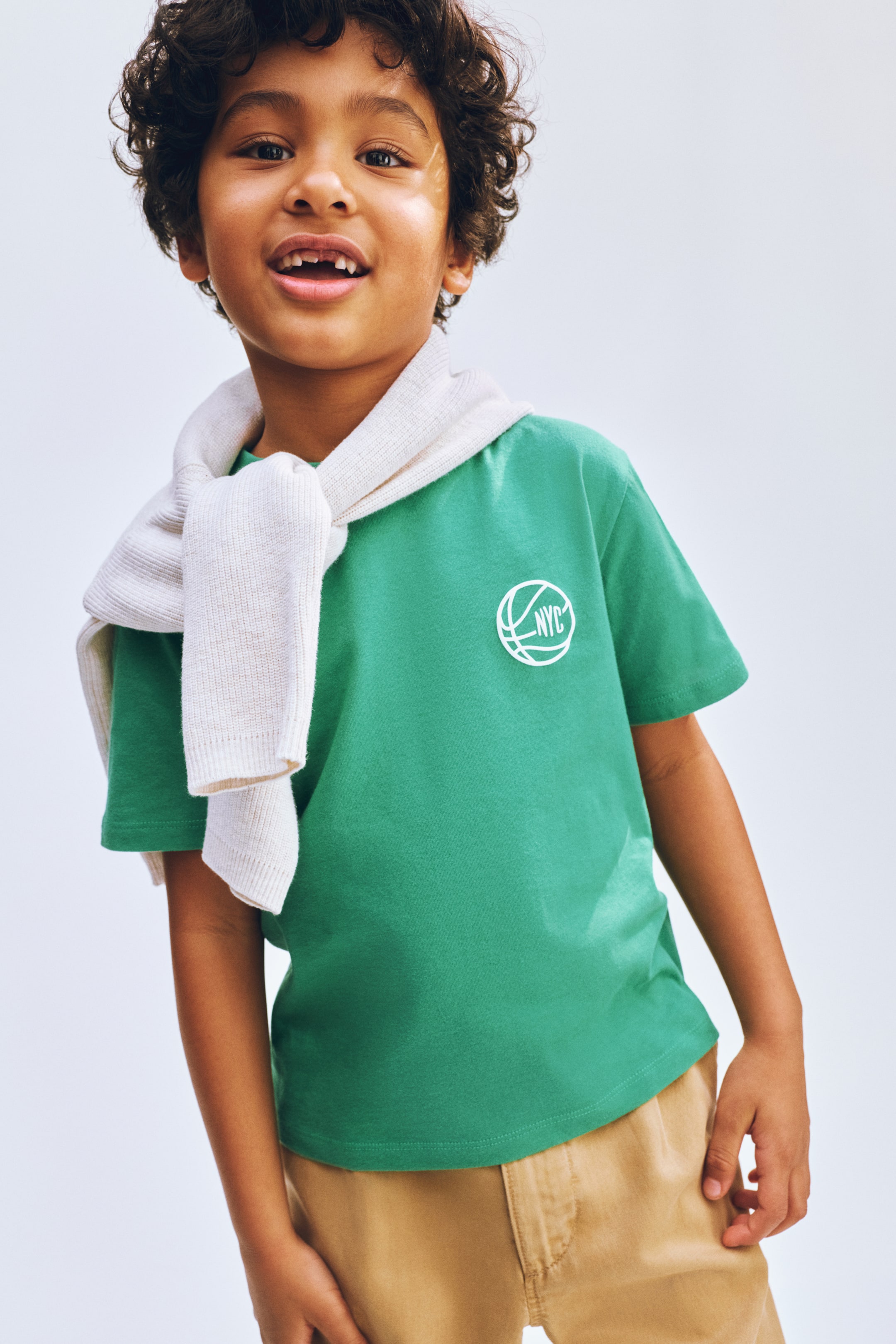 Ver imagem maior: Uma t-shirt verde de manga curta exibe um gráfico circular branco com o contorno de uma bola de basquetebol e "NYC" no seu lado esquerdo do peito. Um casaco de cor clara está pendurado sobre os ombros e o pescoço da t-shirt.