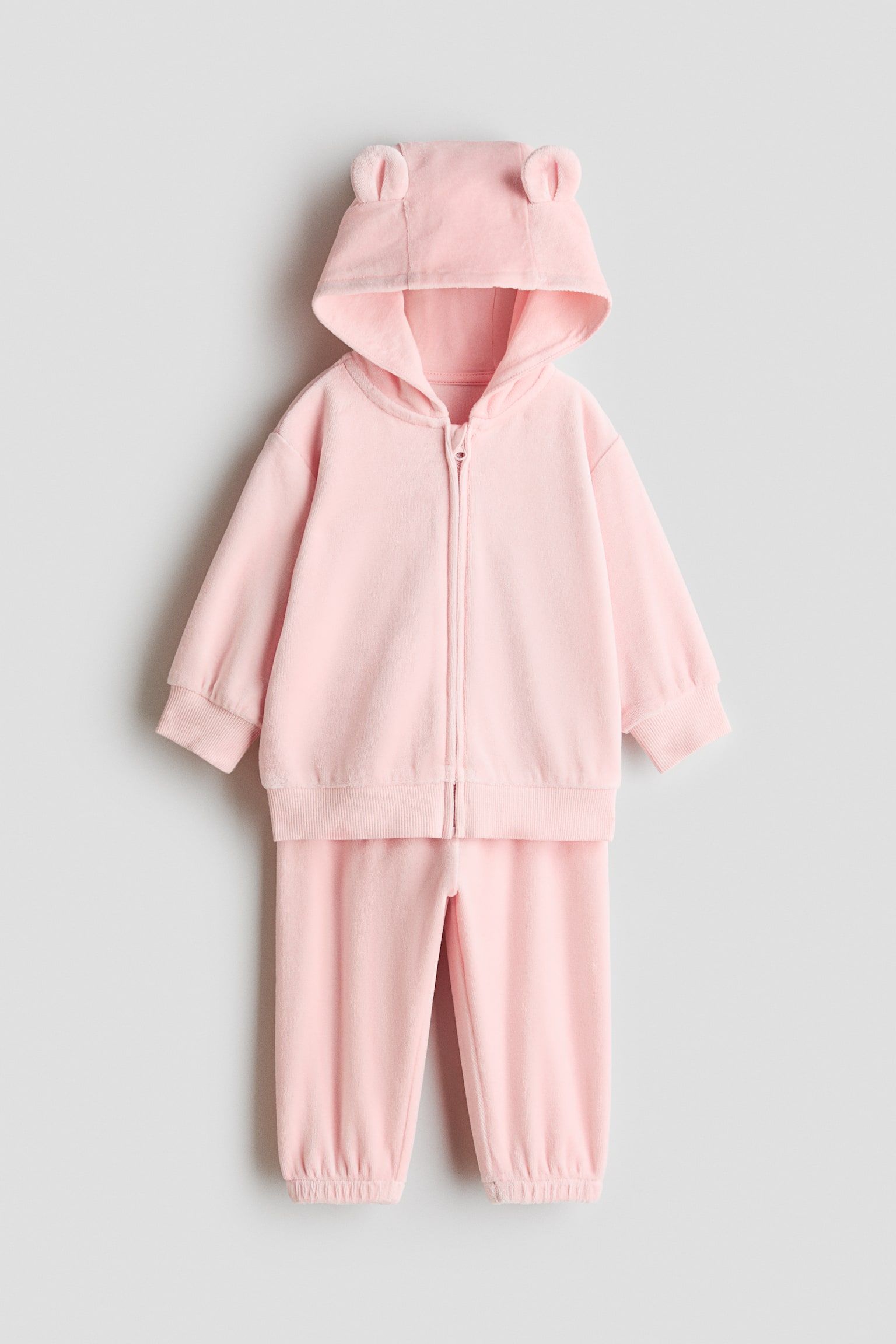 Een lichtroze, fleece-achtig tweedelig setje wordt van voren getoond, bestaande uit een hoodie met ritssluiting en kleine dierenoortjes op de capuchon, geribde boorden en een geribde zoom. De bijpassende broek wordt direct eronder weergegeven en heeft elastische enkelsboorden.