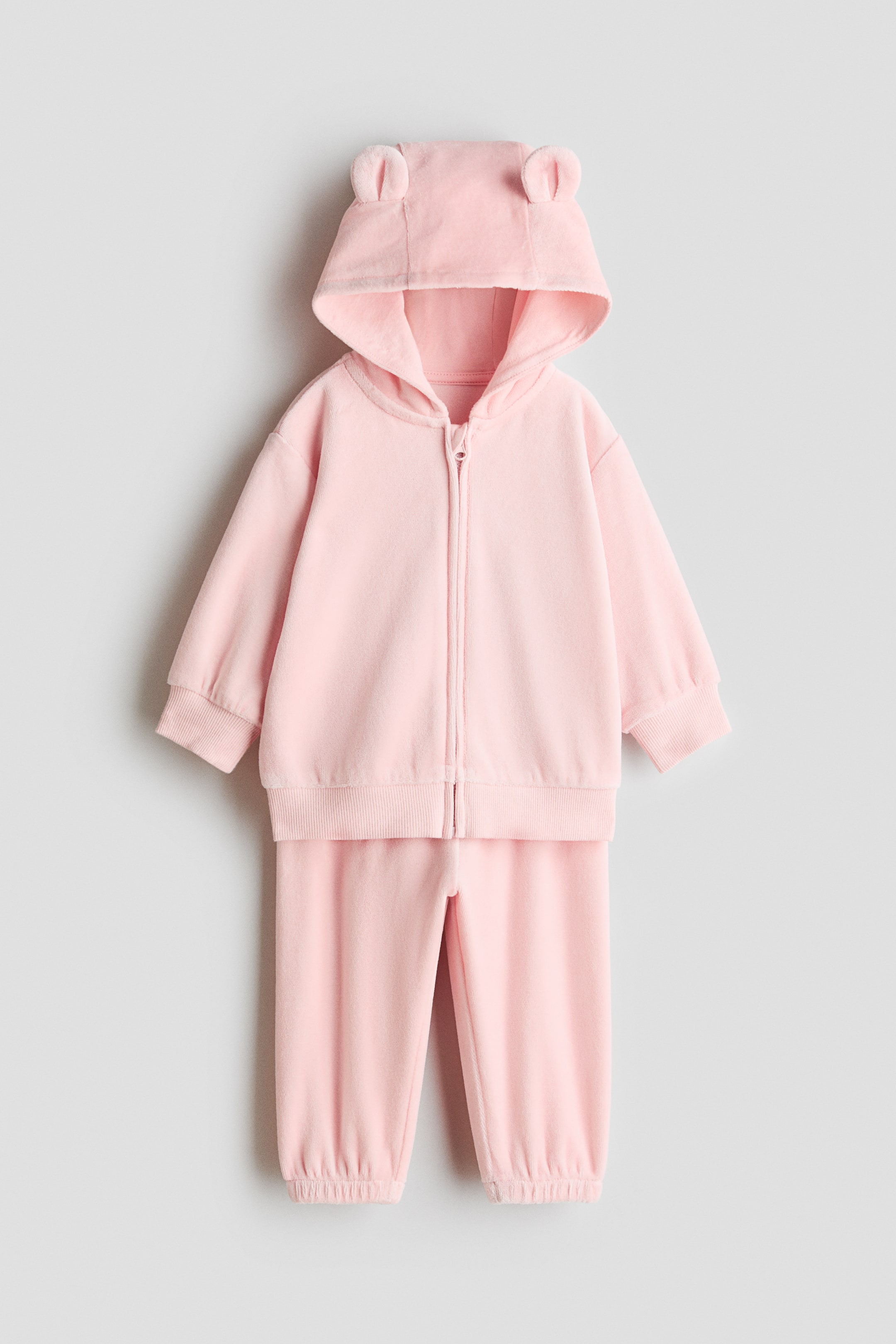 Переглянути збільшене зображення: Світло-рожевий двокомпонентний комплект із матеріалу, схожого на фліс, показано спереду: худі на блискавці з маленькими вушками тваринки на капюшоні, ребристими манжетами та ребристим низом. Відповідні штани розміщені безпосередньо нижче та мають еластичні манжети на щиколотках.