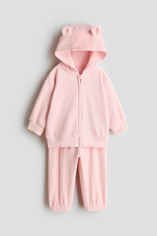 A light pink fleece-like two-piece set is shown from the front, featuring a zip-up hoodie with small animal ears on the hood, ribbed cuffs, and a ribbed hem. The matching trousers are displayed directly below, featuring elasticated ankle cuffs. - 1
