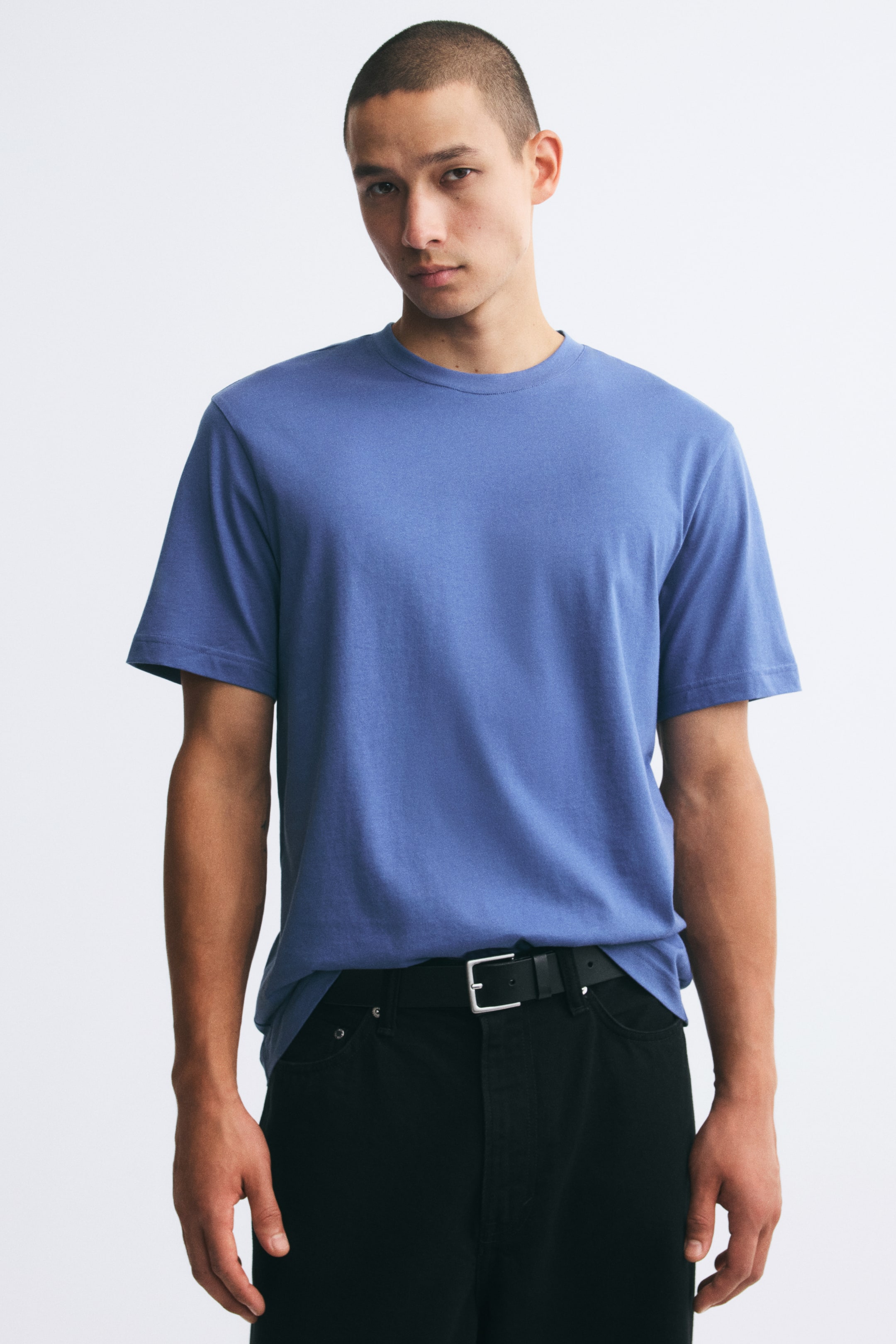 Visa större bild: En blå t-shirt i bomullsjersey med rund halsringning med ribbstickad kant och normal passform bärs av modellen. Tröjan är stylad lätt instoppad i midjan i svarta byxor med ett svart bälte.