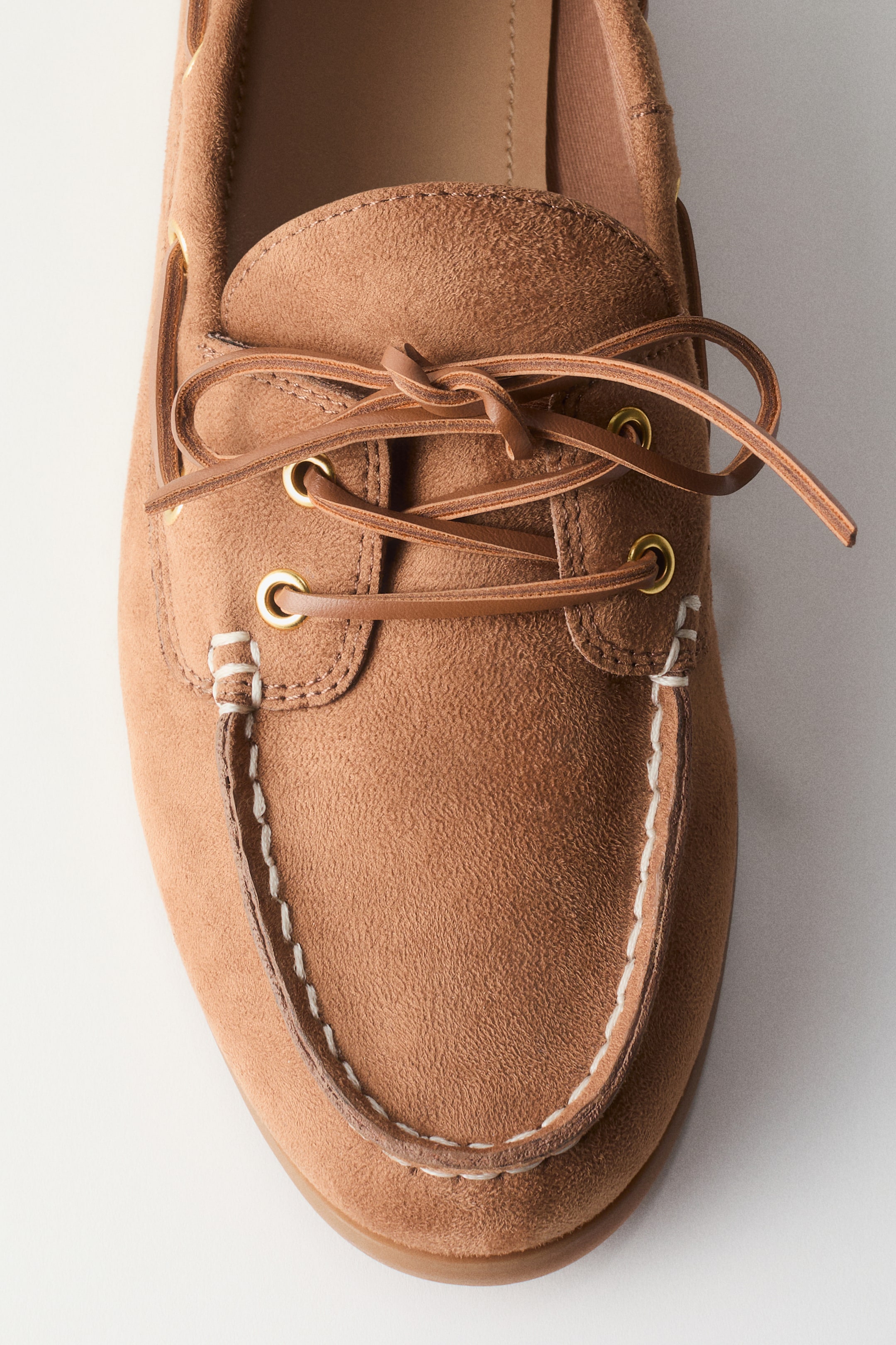 Büyük resmi görüntüle: Kahverengi süet bir tekne ayakkabısı, beyaz mokasen dikişlerini ve kahverengi deri bağcıklarını sergileyerek hafifçe sağa eğimli duruyor. Bağcıklar, altın tonlu perçinlerden geçirilmiş ve ayakkabının üst kısmında bir düğümle bağlanmıştır.