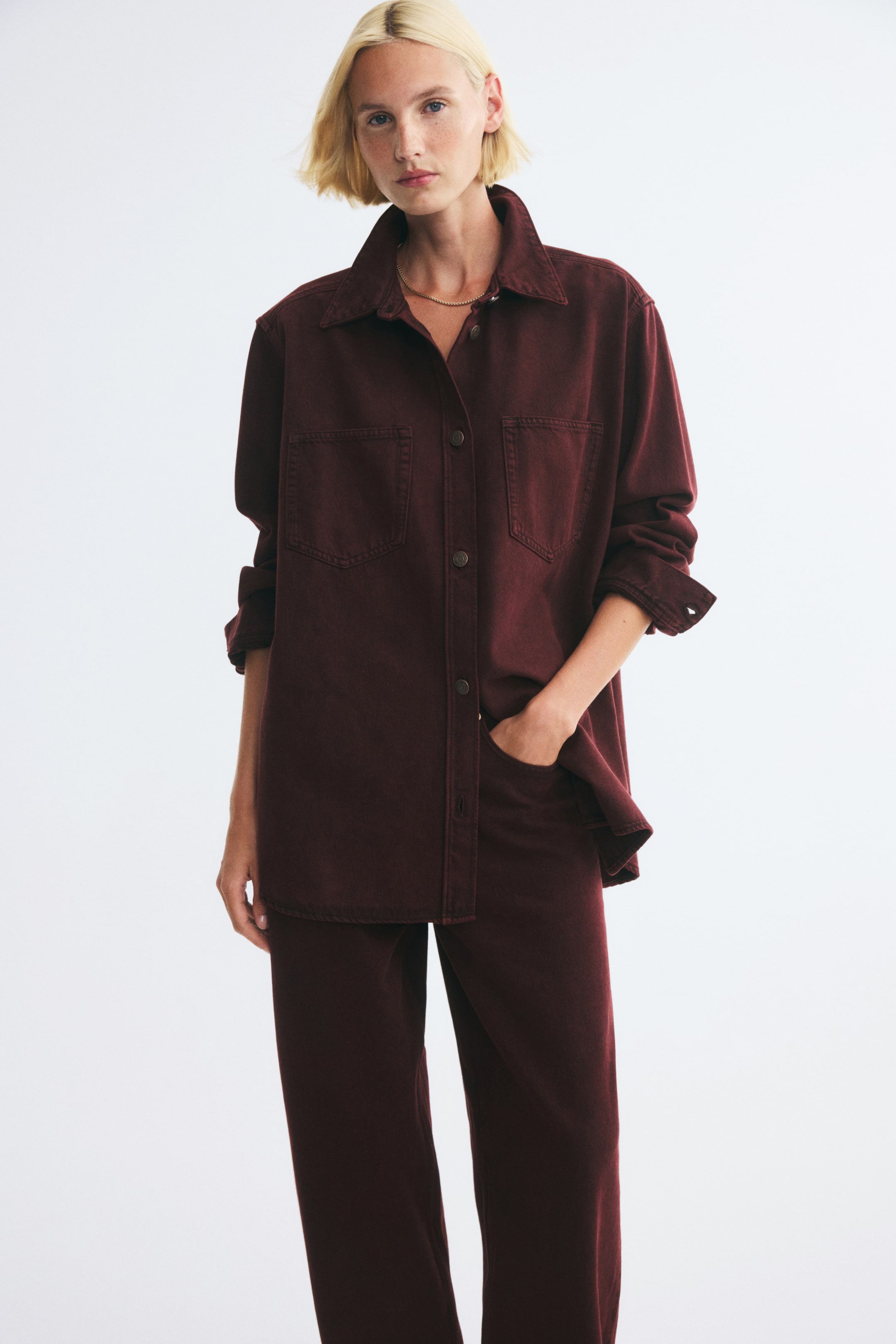 View larger image: An oversized burgundy denim shirt with a collar, button-front closure, and patch chest pockets is worn by the model. The shirt is styled with matching burgundy trousers, with the model placing one hand in a trouser pocket.
