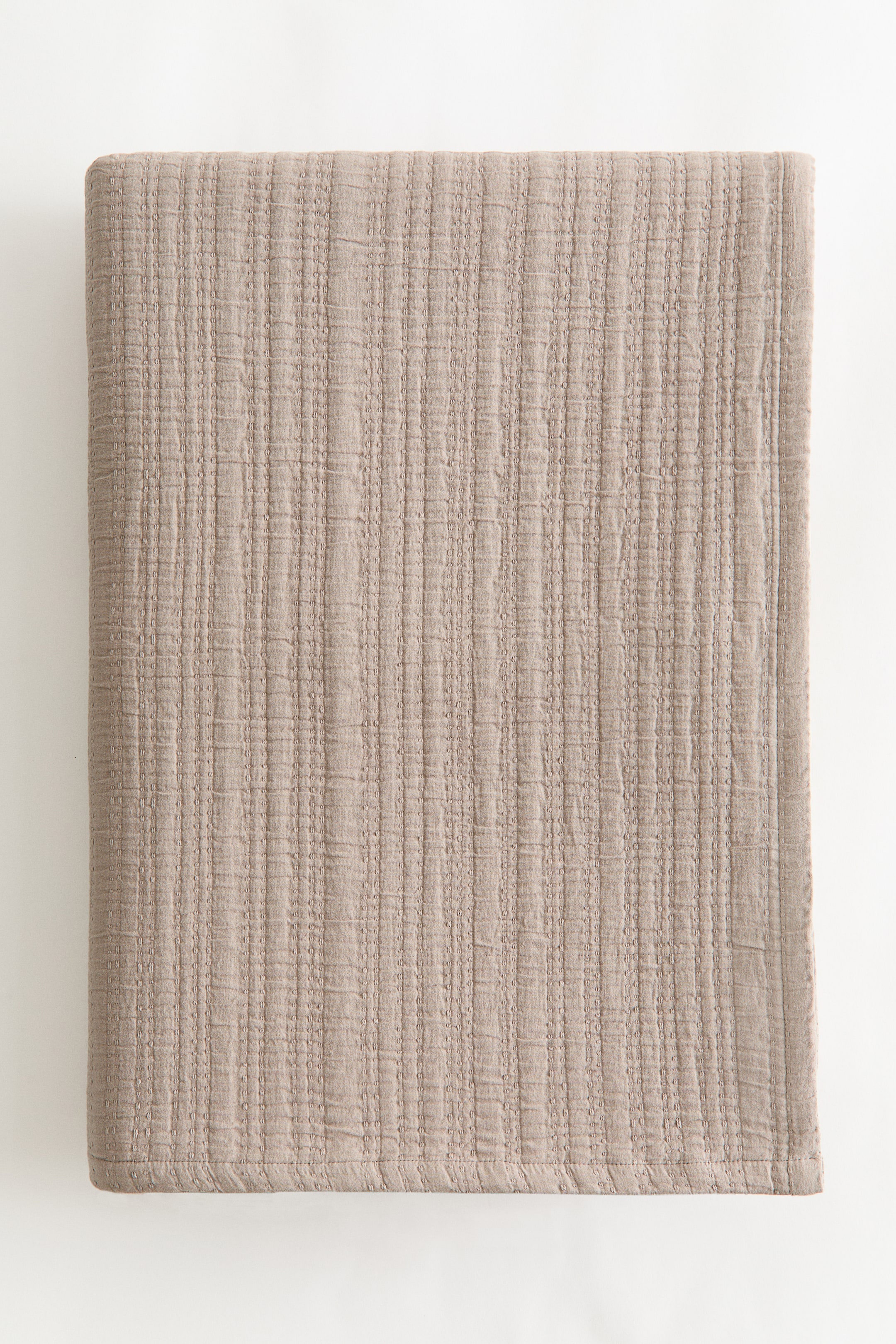 View larger image: A double bedspread in a soft cotton muslin weave is shown from the front, folded flat. It is displaying a beige colour and a textured surface with distinct vertical stitched lines.