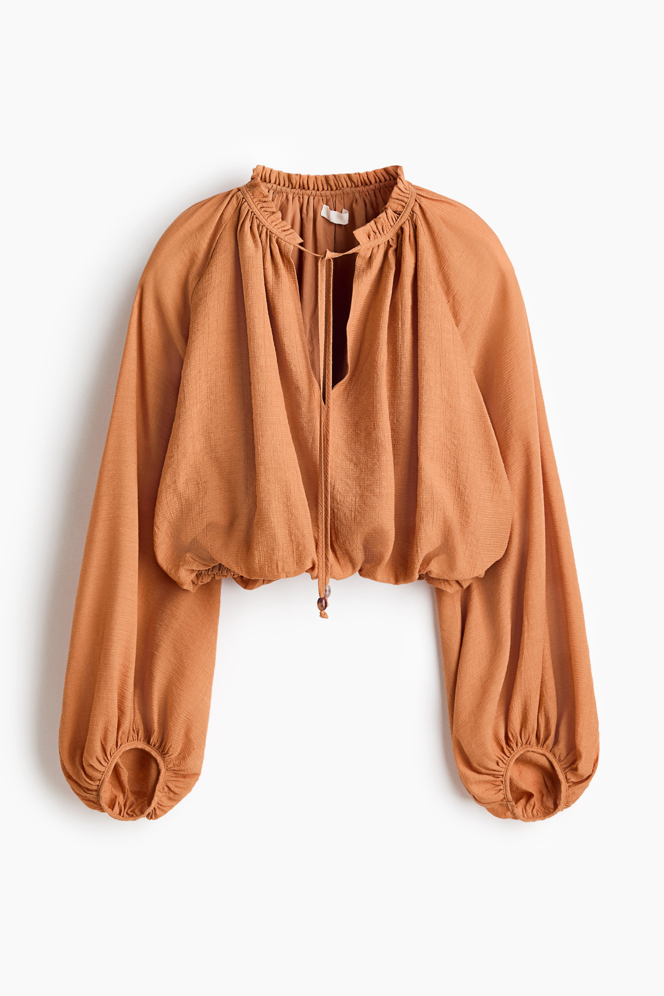 View larger image: A rust-hued cropped blouse exhibits a ruffled high neck with ties at a V-split opening. It showcases voluminous raglan sleeves with gathered cuffs and an elasticated hem adorned with a drawstring and wooden beads.