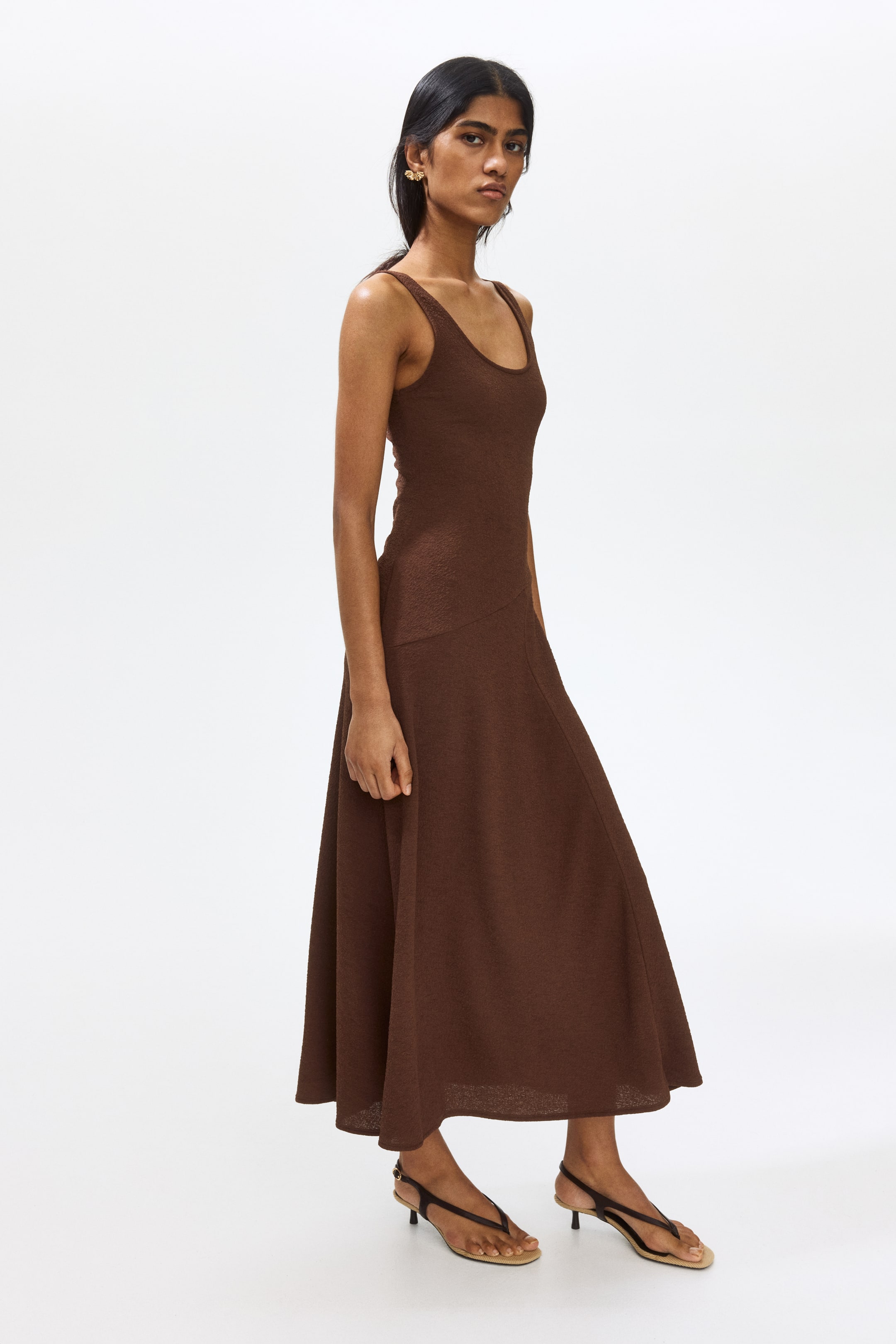 View larger image: A brown midi dress featuring a scoop neckline and tank top straps. It showcases a fitted bodice that transitions into a flared skirt with a subtle textured knit fabric, presented from a slight side angle.