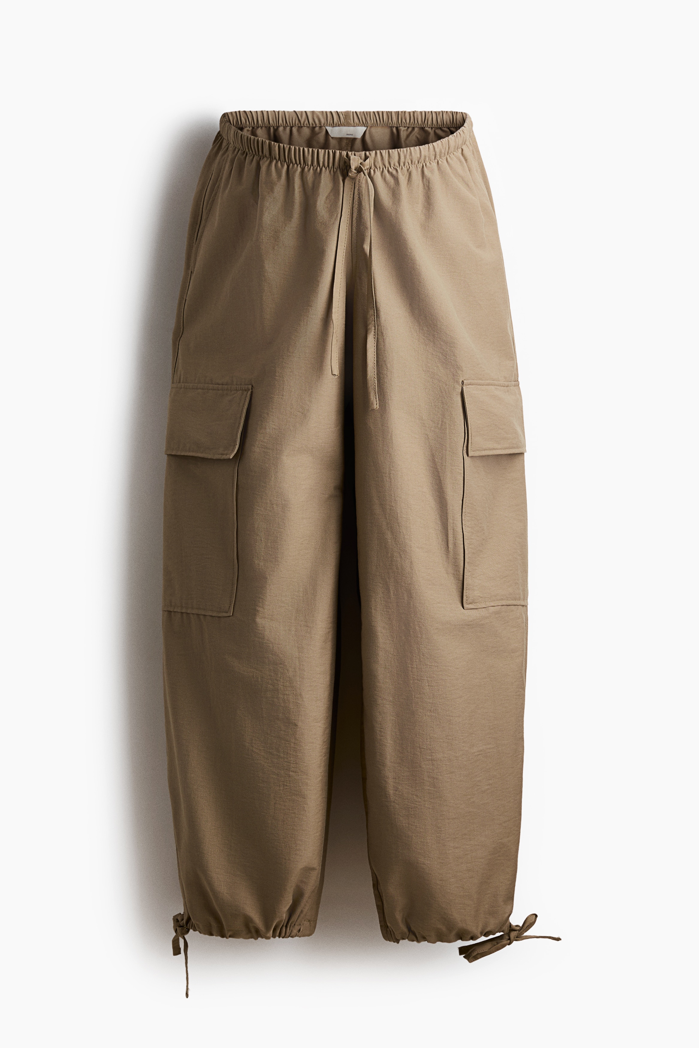 The model is wearing relaxed-fit cargo trousers in a tan cotton blend, styled with a long-sleeve cream top. The trousers feature a low, elasticated waistband with a tied drawstring, side cargo pockets, and balloon legs with adjustable drawstrings at the ankles.