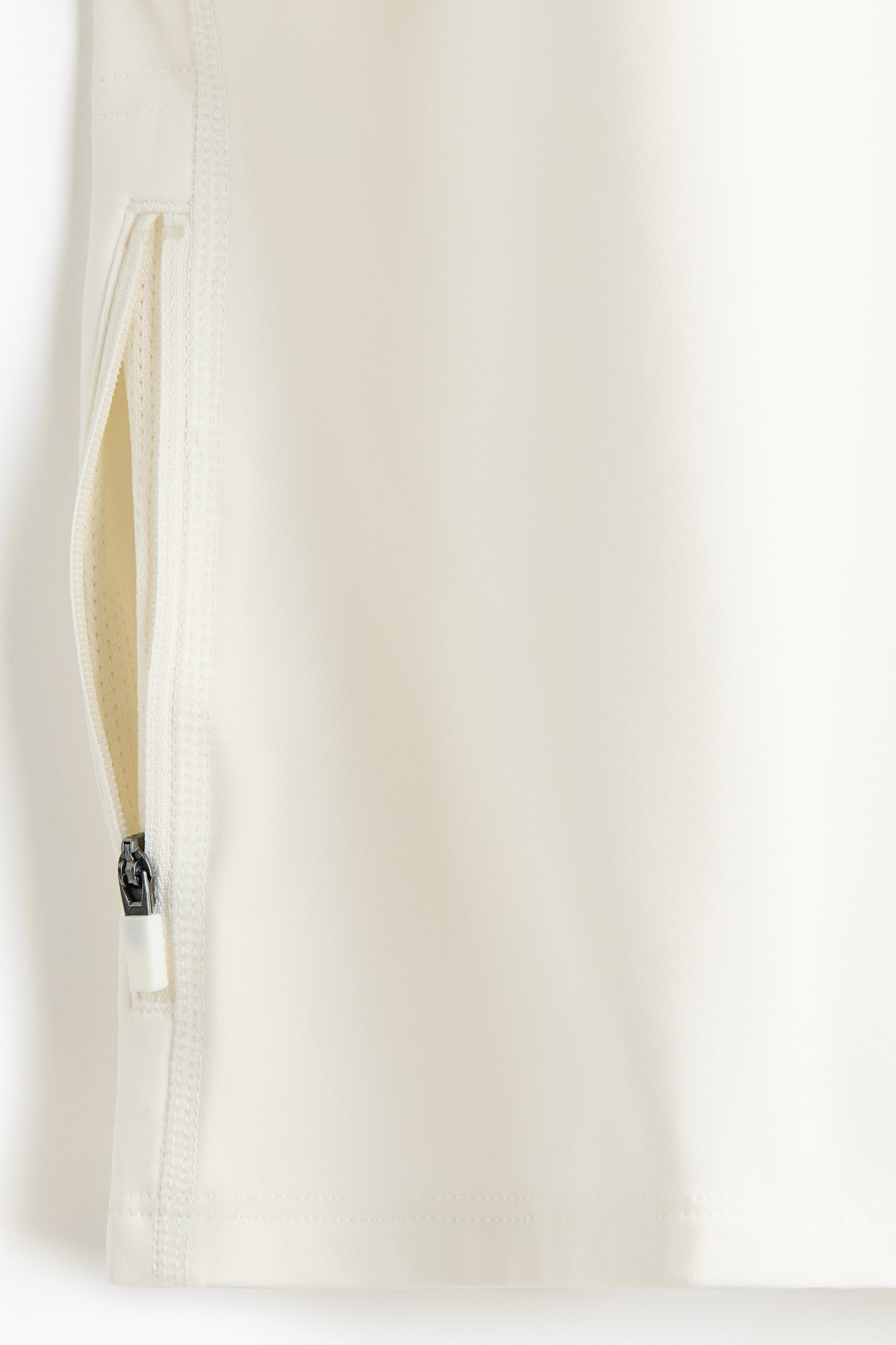 Ampliar la imagen: Un primer plano de una prenda de color blanco hueso muestra un bolsillo con cierre parcialmente abierto en su lado izquierdo. El bolsillo revela un forro de malla de color claro, y un tirador de cierre oscuro con una pestaña blanca es visible en la parte inferior de la abertura, con pespuntes visibles a lo largo del cierre y el dobladillo de la prenda.