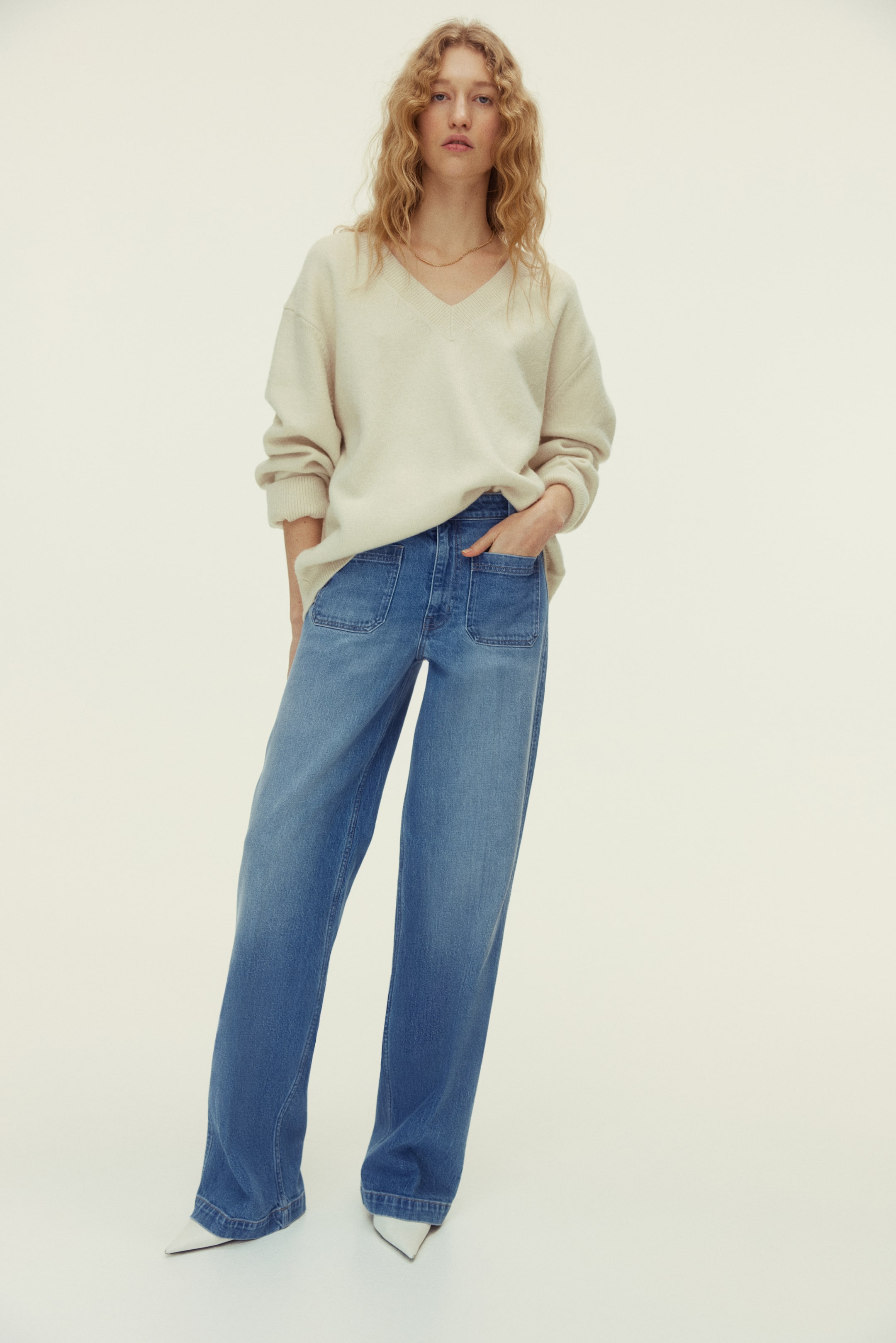 View larger image: An off-white, relaxed-fit V-neck sweater featuring long sleeves and a soft texture. It is partially tucked into blue, wide-leg denim jeans designed with prominent front patch pockets.