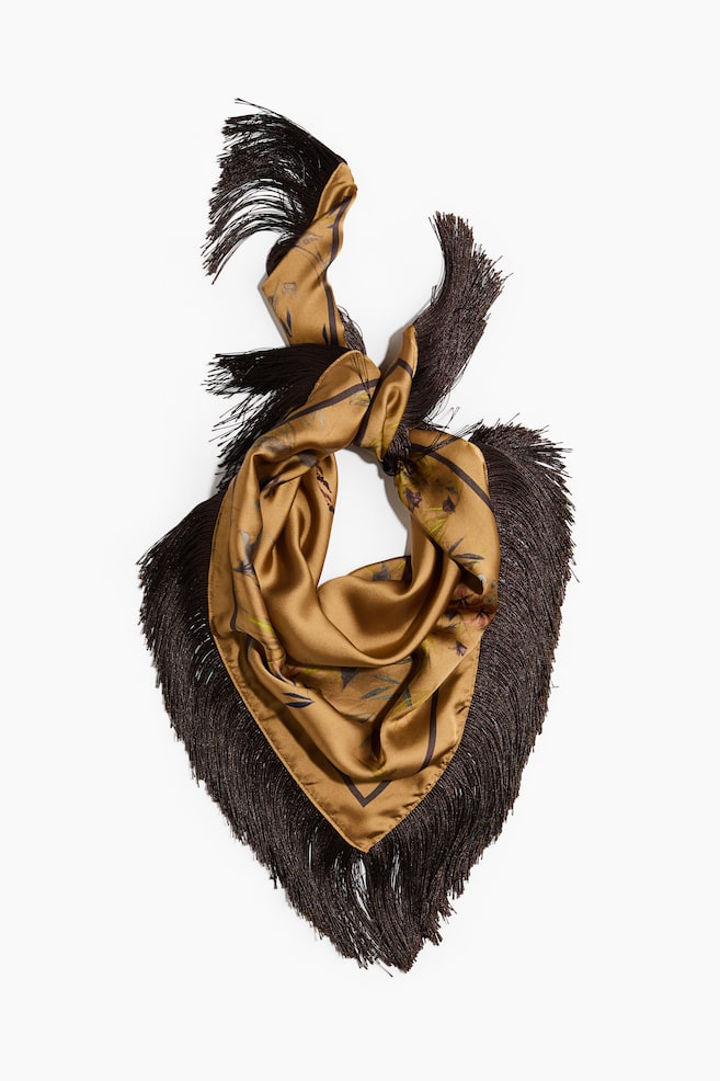 A golden-brown silk scarf featuring a subtle botanical print is displayed from the front, loosely knotted at the top to form a draped triangular shape. Long, dark brown fringe borders the edges, cascading downward and outward. - 1